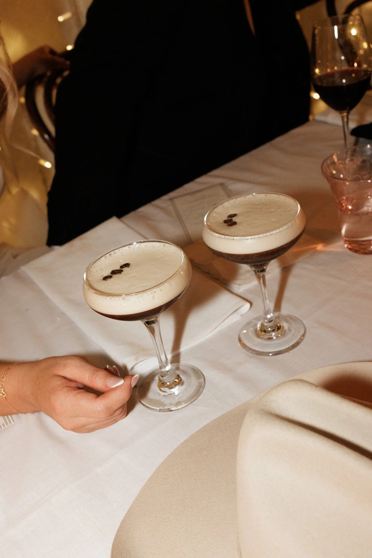 A person's hand holding three cocktail glasses with foamy dark drinks and coffee beans on top, on a white table with other glasses and a person in black sitting nearby.