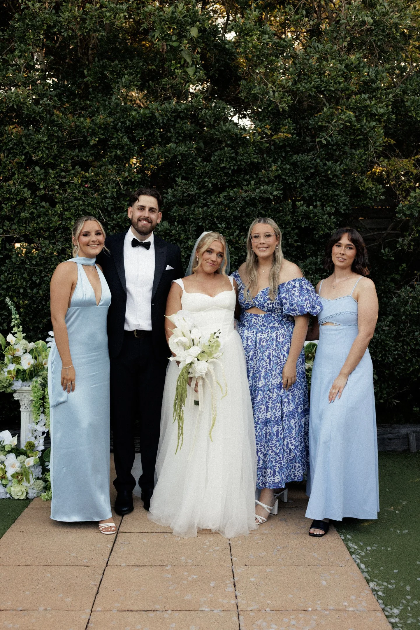 Group of five people dressed in formal clothing, standing outdoors in front of a large green bush, celebrating a wedding. The bride wears a white gown holding a bouquet, surrounded by four women in pastel-colored dresses and a man in a tuxedo.