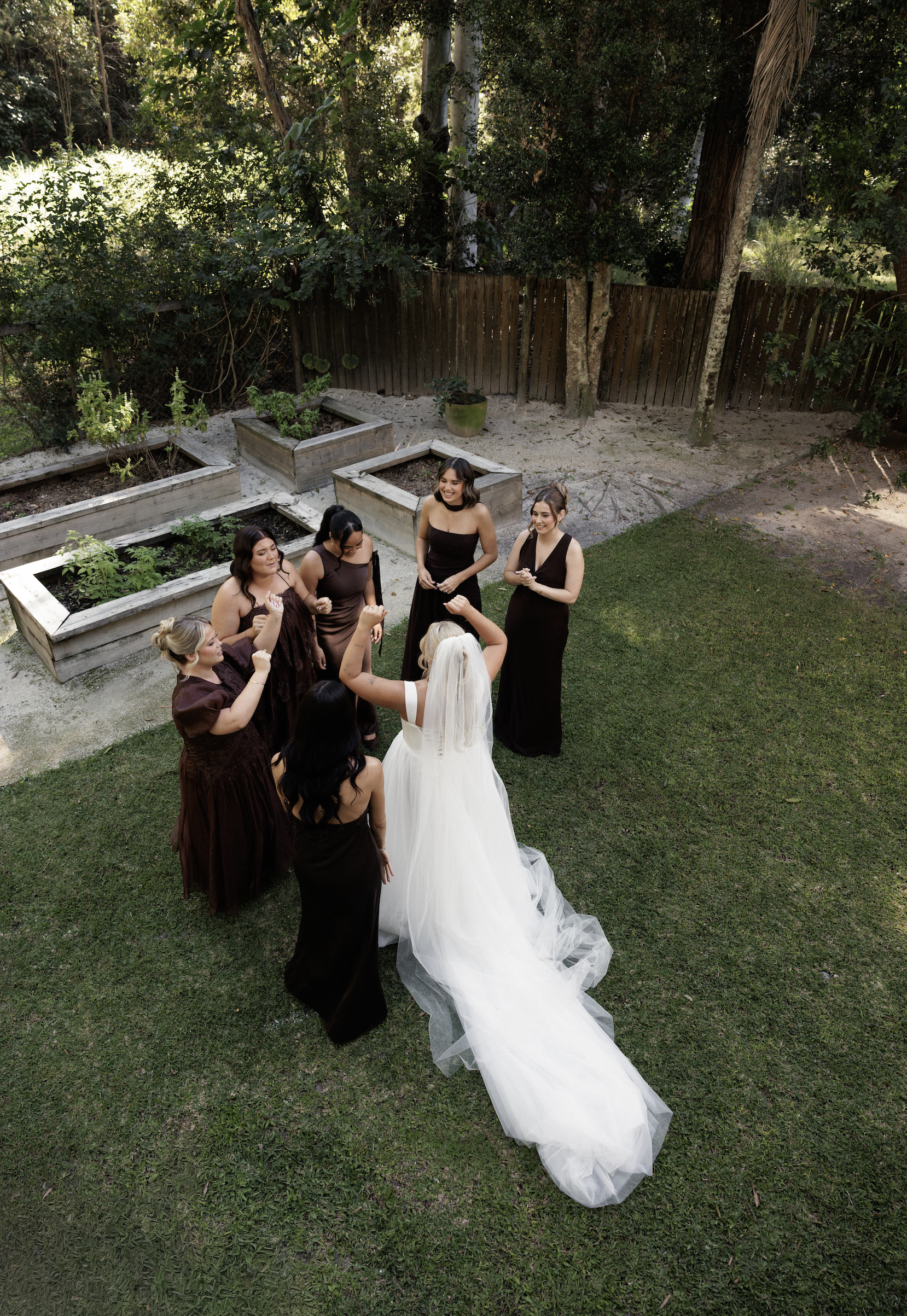 A group of women in dark dresses and a bride in a white wedding gown gather outdoors, celebrating together.