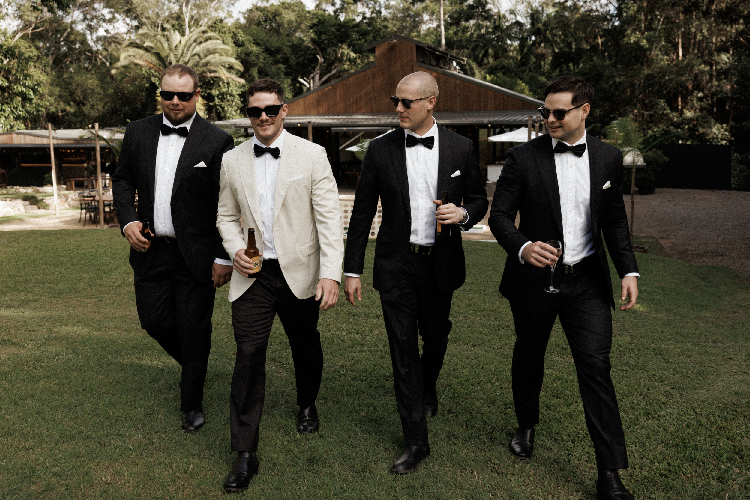 Four men dressed in tuxedos, wearing sunglasses, walking on a lawn holding drinks, with a wooden building and trees in the background.