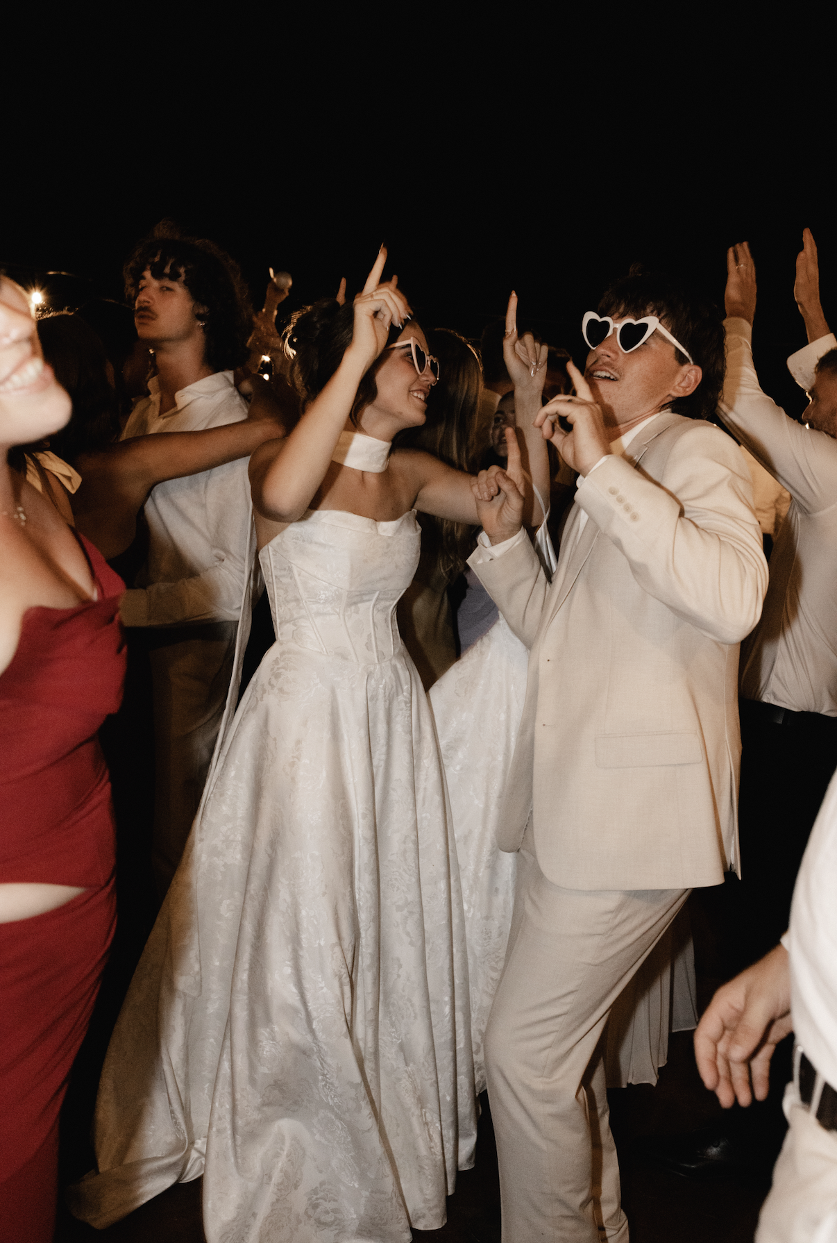 People dancing at a wedding reception at night, wearing stylish formal attire, with some individuals wearing playful sunglasses.