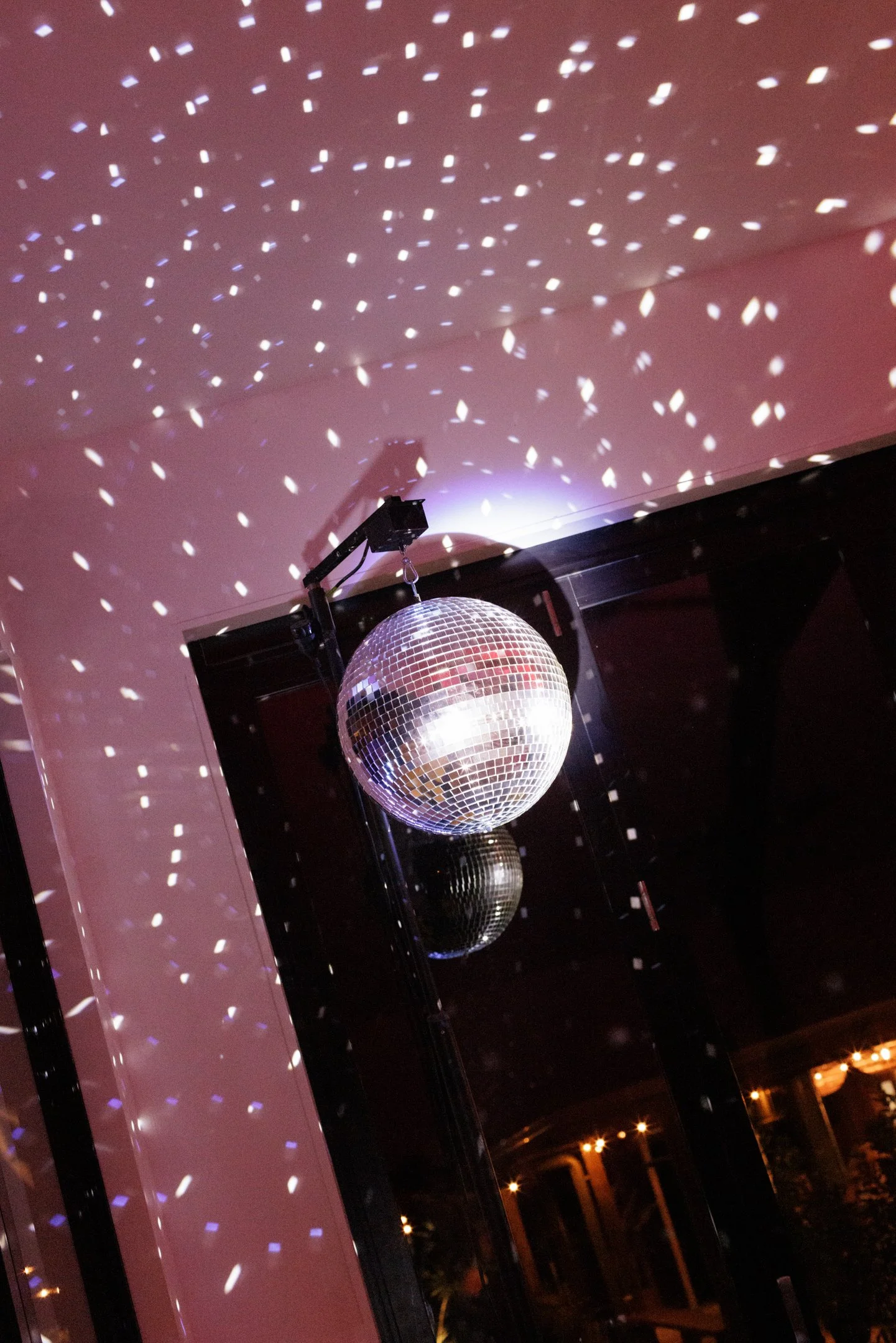 A disco ball hanging from the ceiling with light reflections creating a pattern on the pink wall and ceiling in a dark room.