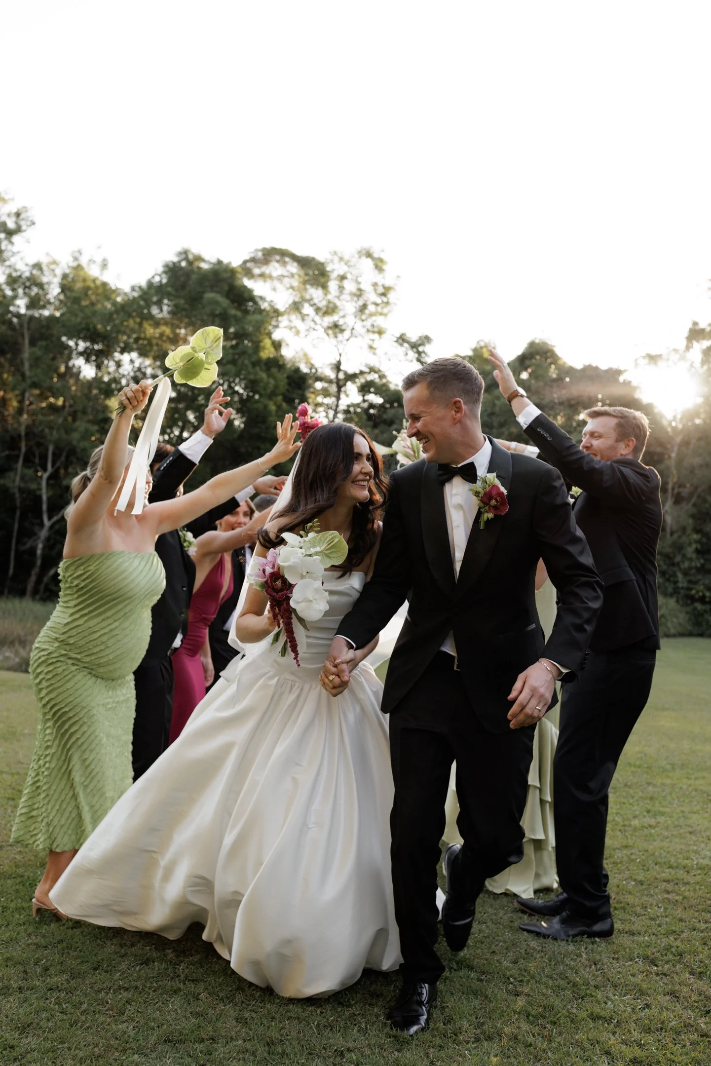 A bride and groom holding hands and smiling at each other as they walk outside. The bride is wearing a white wedding gown and veil, holding a bouquet of white and burgundy flowers. Guests surround them, tossing green leaves and flower petals. The scene is set in a grassy area with trees and a bright sky in the background.
