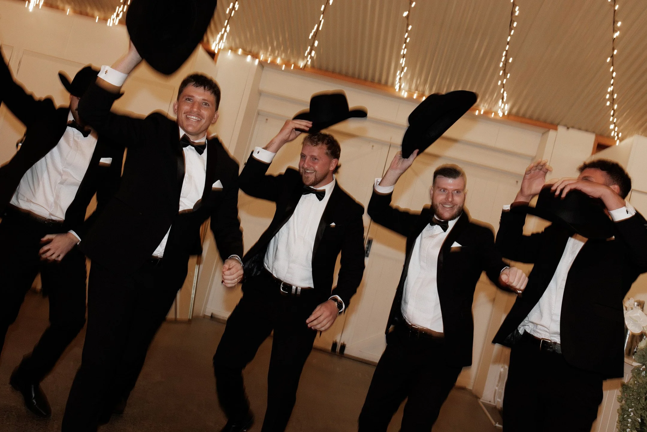 Group of men dressed in tuxedos celebrating at a party, some holding black hats, with string lights hanging from the ceiling in the background.