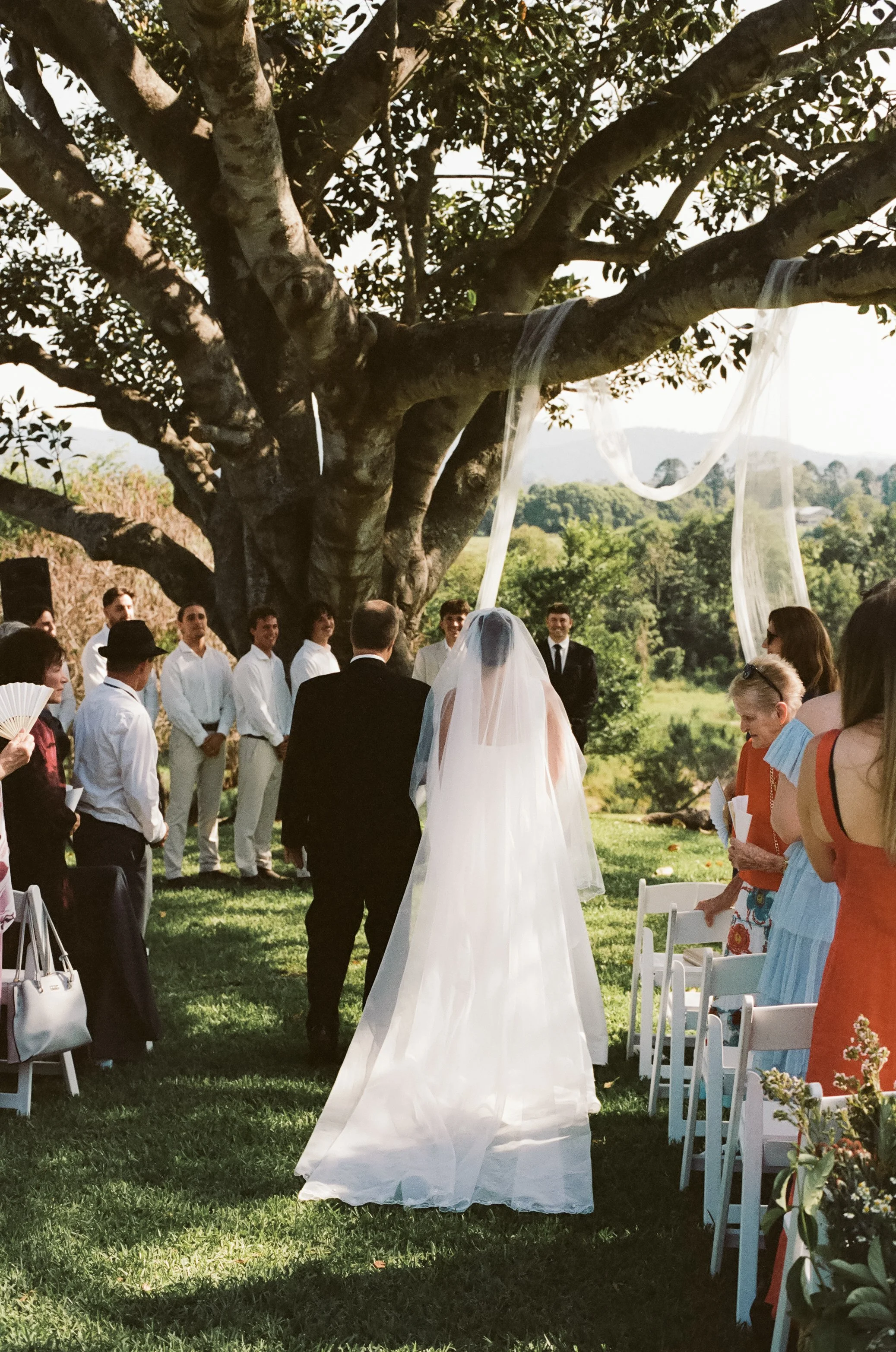 Bride and groom standing under a tree during an outdoor wedding ceremony, with friends and family gathered around in a lush green landscape.