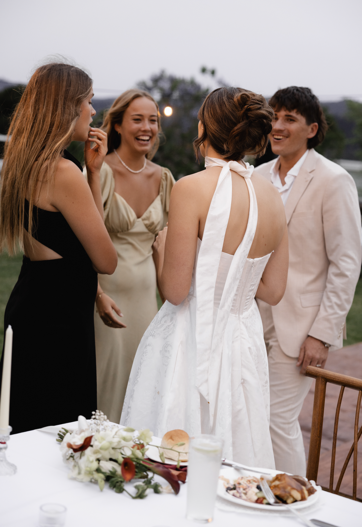 Group of four people at an outdoor wedding reception, with a woman in a white wedding dress talking to three friends.