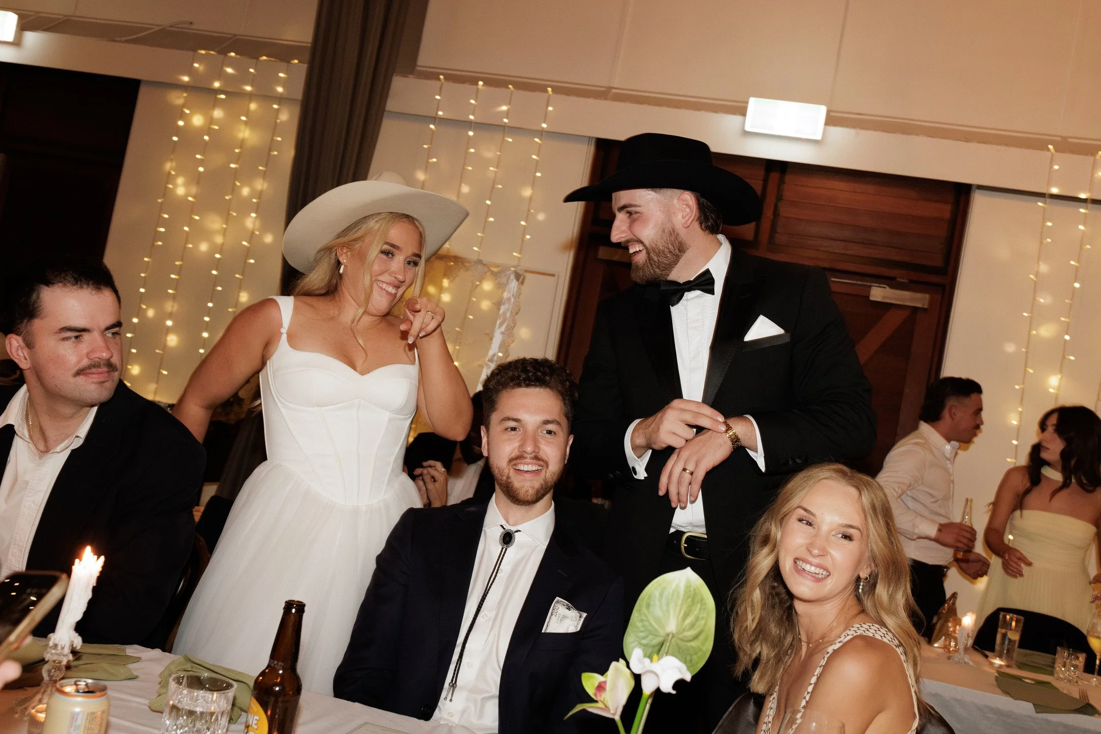 People celebrating at a wedding reception with string lights and floral decorations in the background.