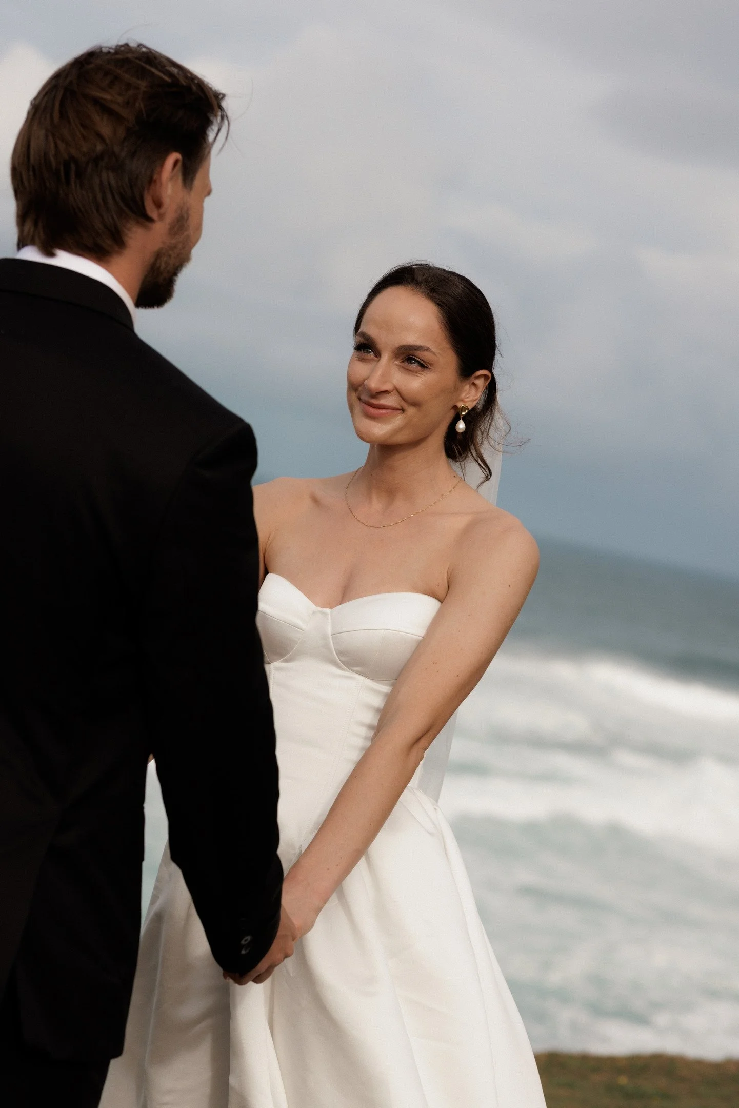 A couple dressed in wedding attire holding hands by the seaside, with the woman smiling and looking at the man, in front of a cloudy sky and ocean.