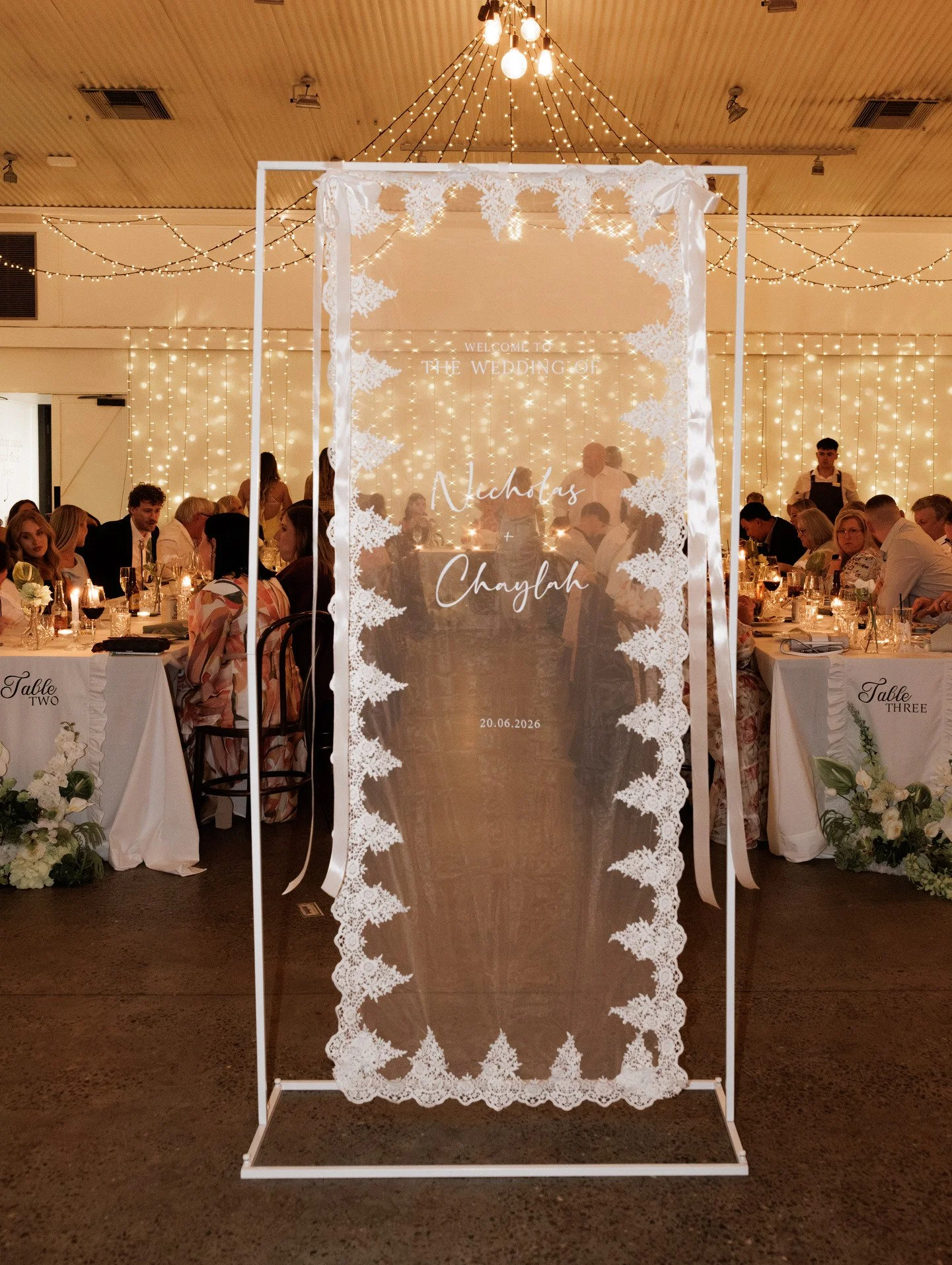 Wedding reception with guests seated at decorated tables, fairy lights hanging from ceiling, and a decorative fabric panel with lace patterns and the names 'Nicholas' and 'Chaylah' along with the date '20.06.2026' in the foreground.