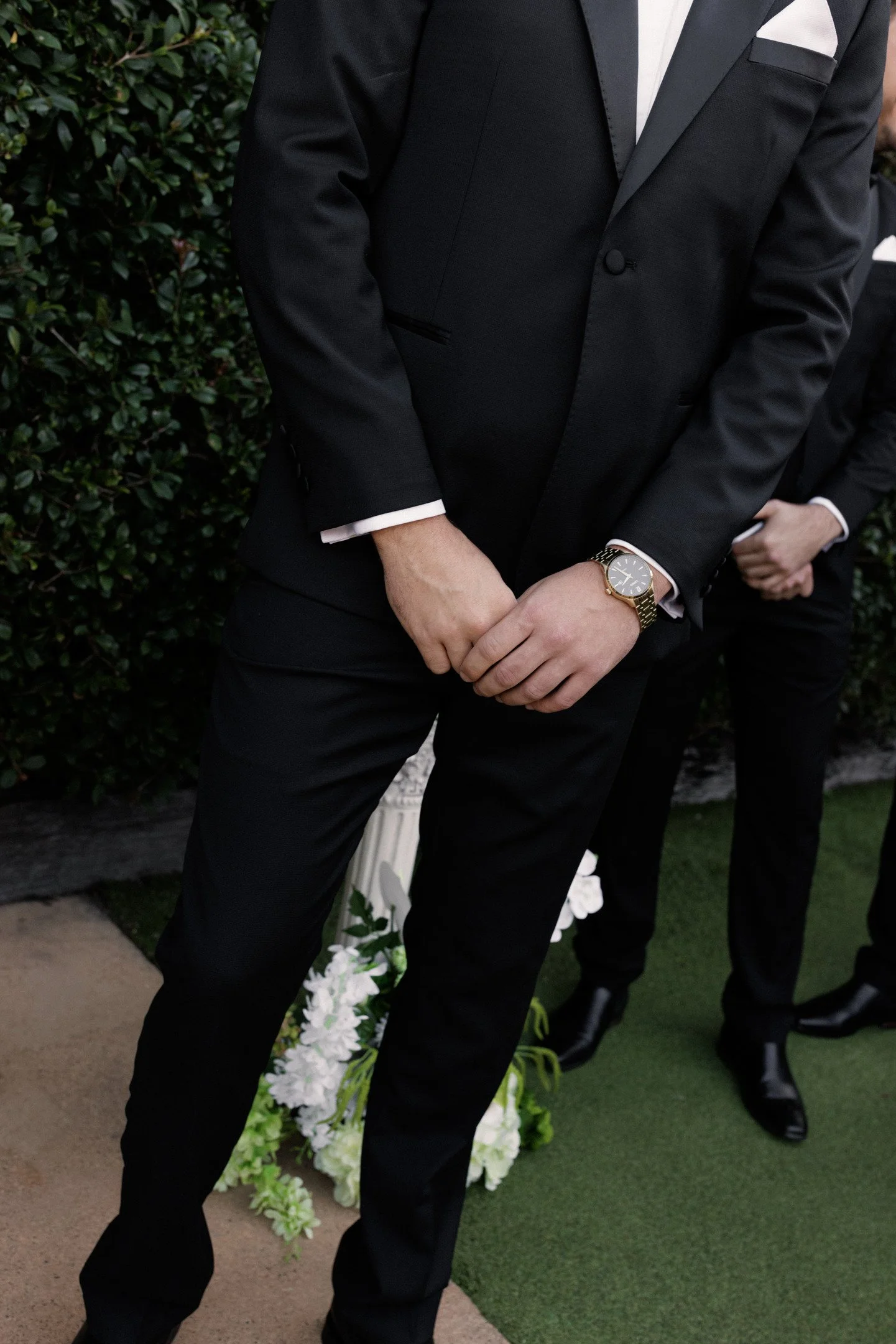 A person dressed in a black suit with a white shirt and a gold wristwatch, standing outdoors near a green hedge and floral arrangement.