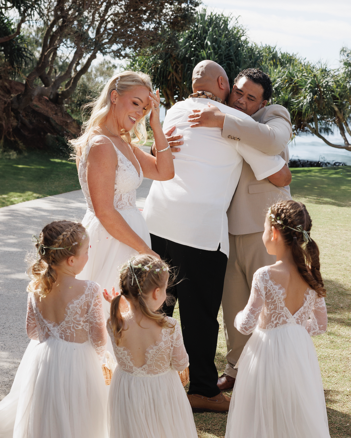A wedding scene outdoors with a bride, groom, and two men hugging, surrounded by three young girls in white dresses with lace tops and floral headbands.