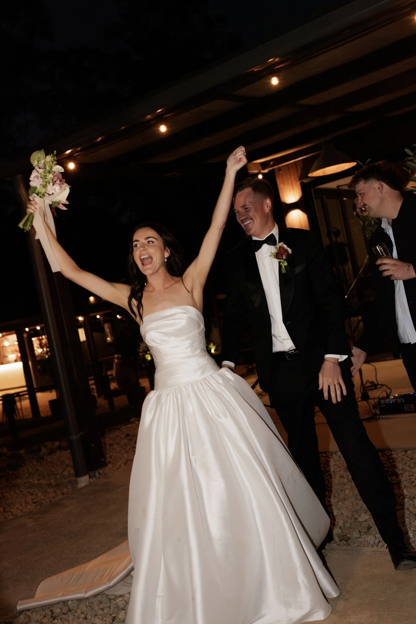 A bride in a white wedding dress celebrating with two men dressed in tuxedos at an indoor wedding reception, with warm lighting and decorative elements in the background.