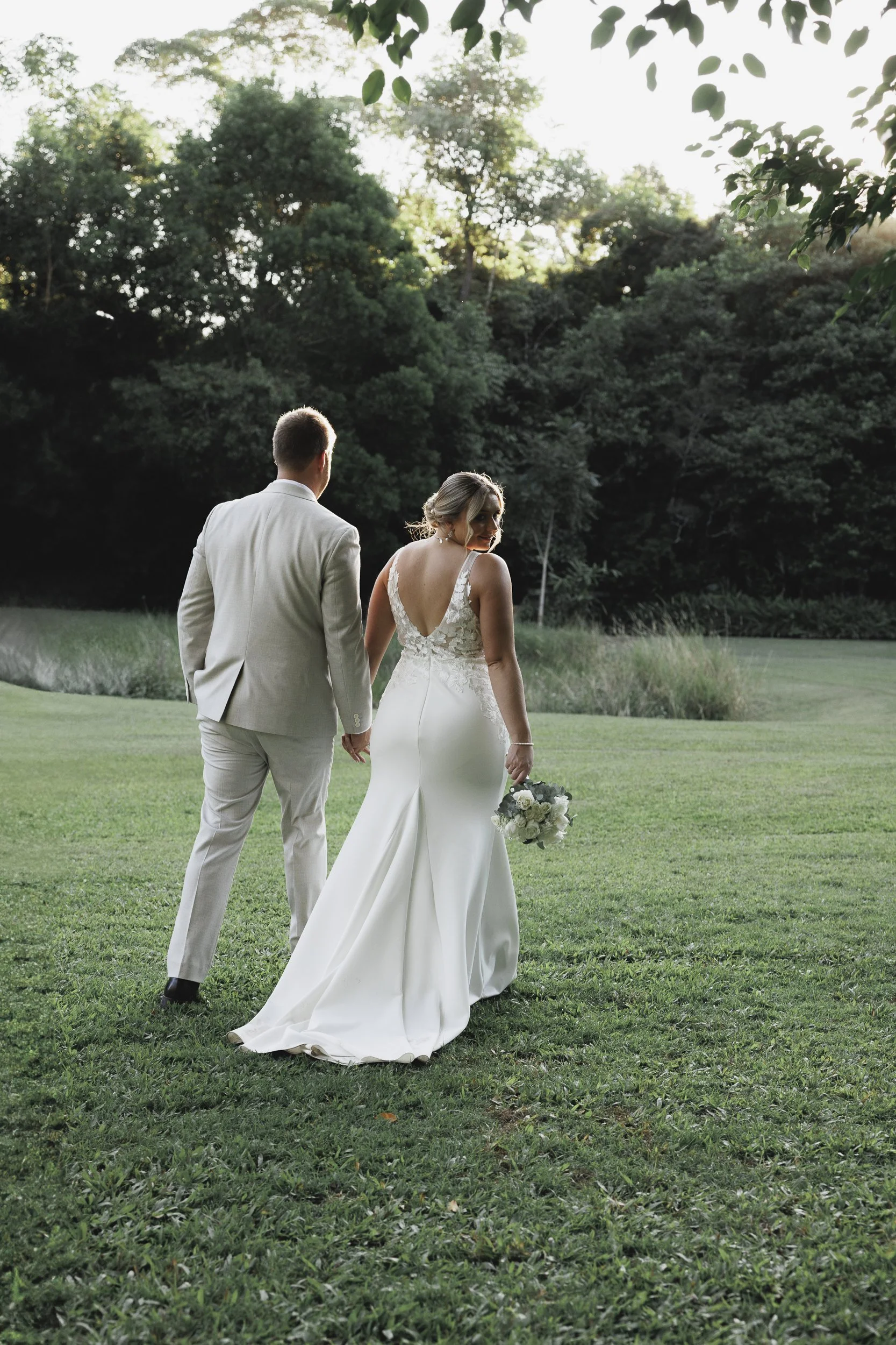 Bride and groom walking hand in hand on a grassy field, with the bride holding a bouquet of white flowers, surrounded by trees and greenery during a wedding photoshoot.