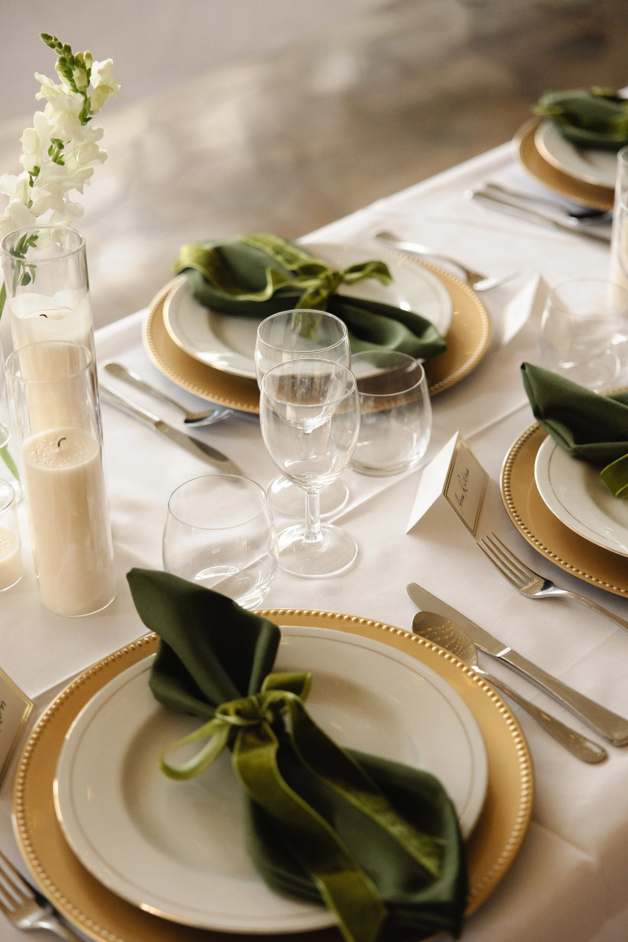 Elegant table setting with white plates on gold chargers, green napkins, glassware, candles, and a white flower centerpiece Elevated Weddings