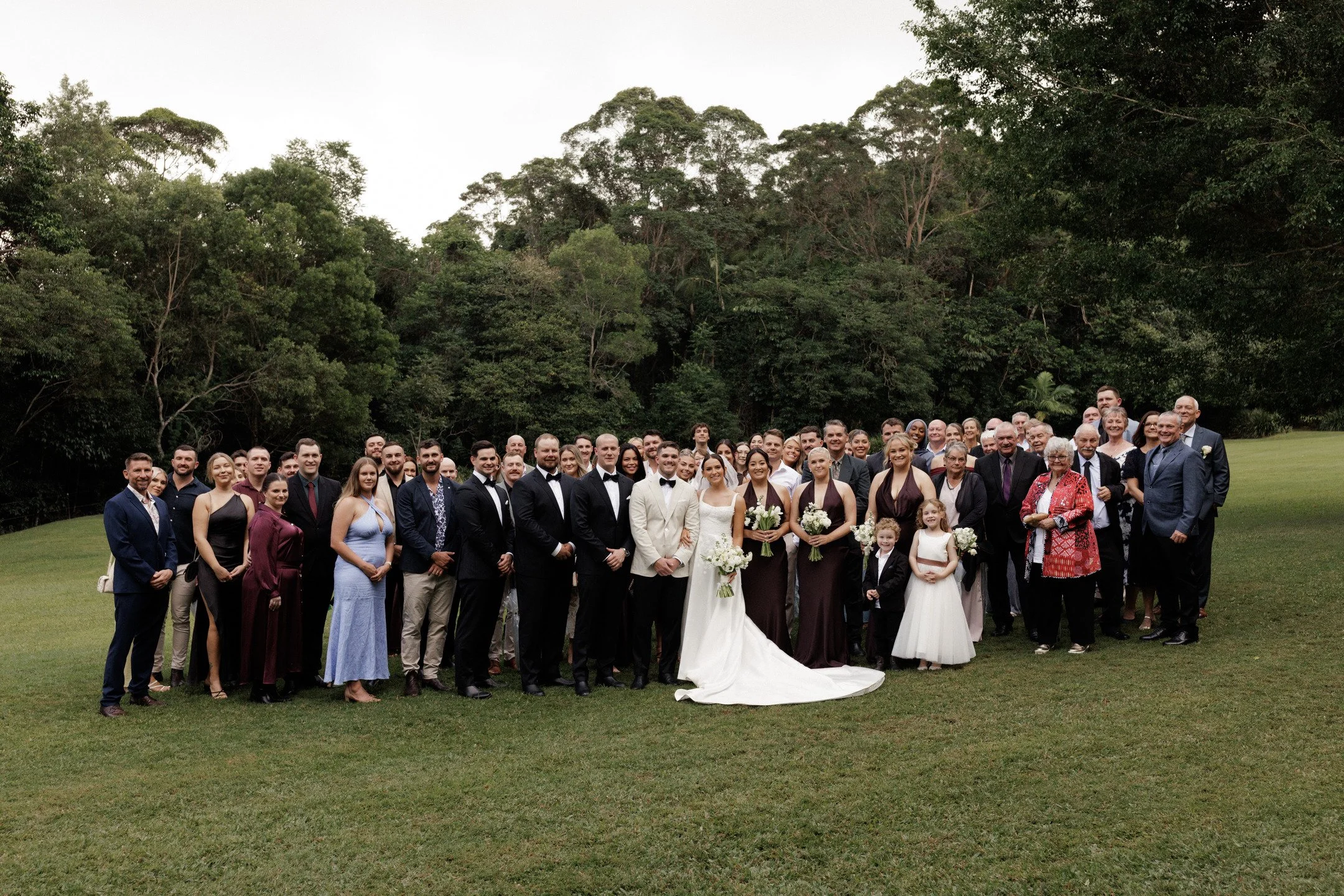 Large group of people gathered outdoors on grass with dense trees in the background, including men, women, and children dressed formally, some in wedding attire.