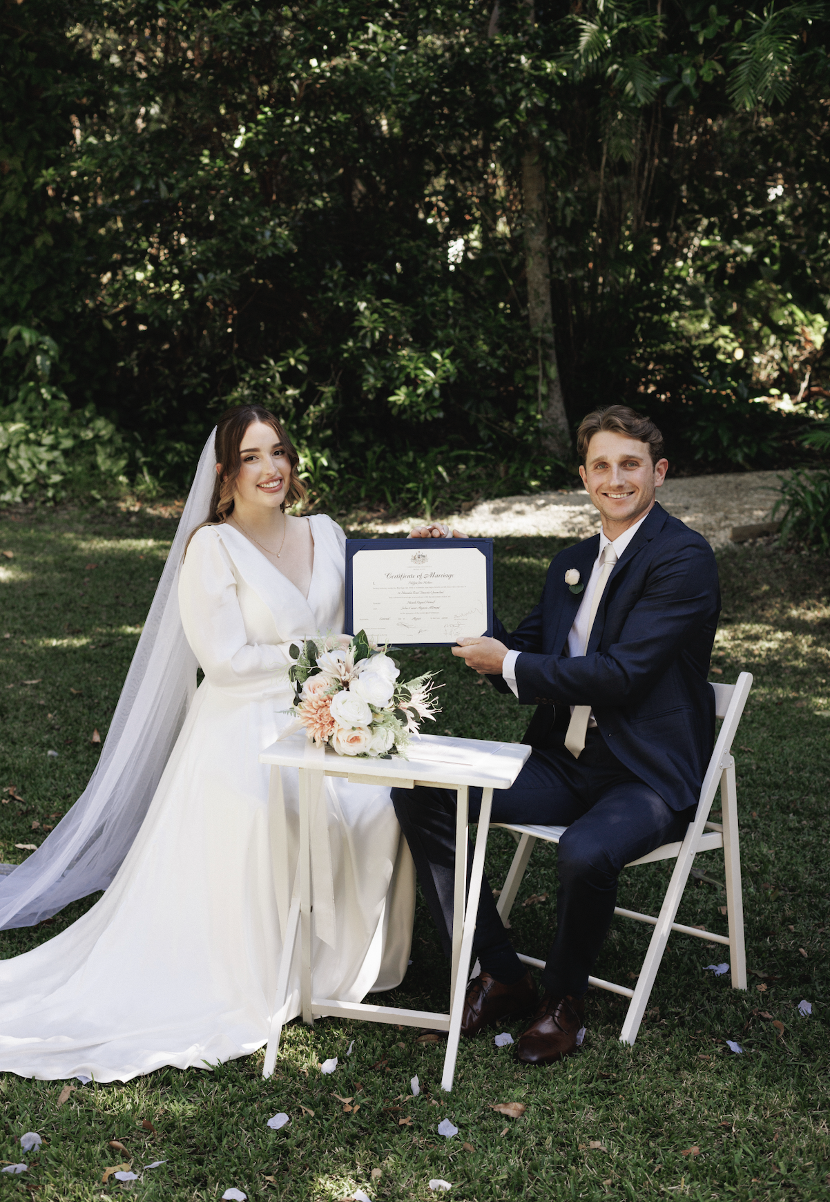 A bride and groom sit outdoors on a lawn, holding a marriage certificate together, smiling. The bride wears a white wedding dress with a veil, and the groom is in a navy suit with a white shirt and light-colored tie. A bouquet of white and blush pink