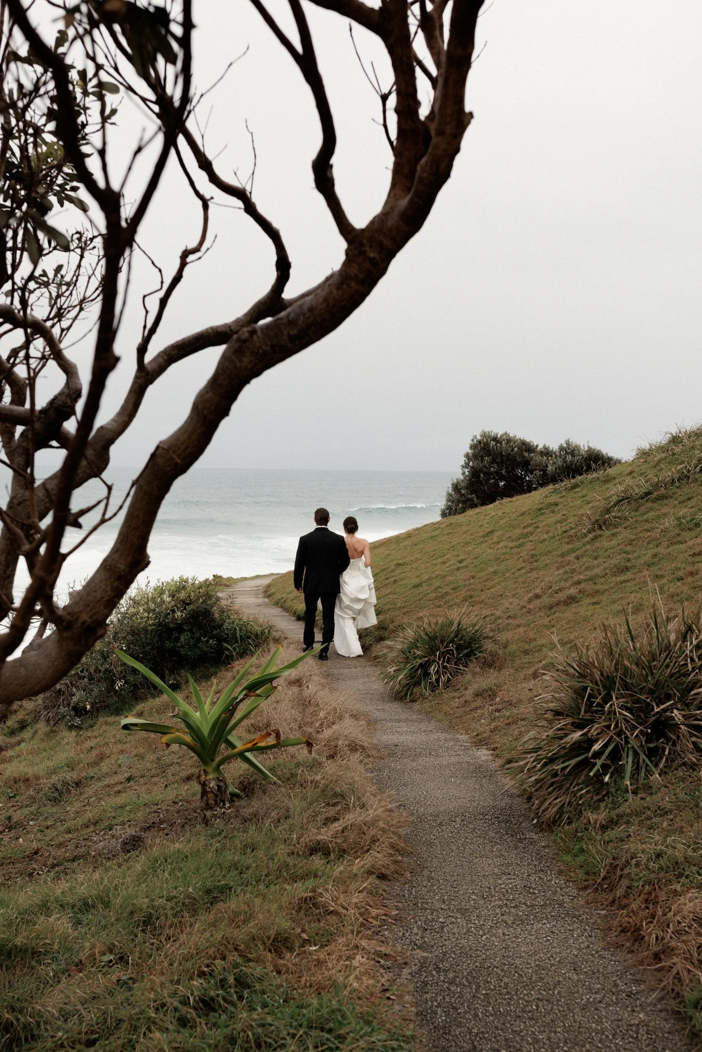A couple dressed in wedding attire walking down a narrow beachside path surrounded by green plants, with the ocean in the background on a cloudy day.