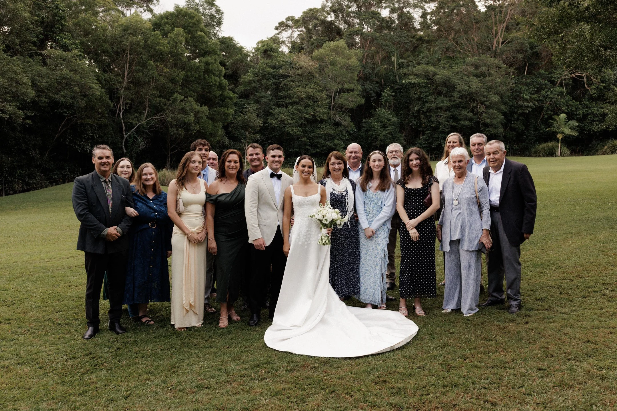 A large group of people, including a bride and groom, poses outdoors on a grassy field with a backdrop of trees. The bride is in a white wedding dress holding a bouquet, and the groom is in a light-colored suit with a black bow tie. The group appears