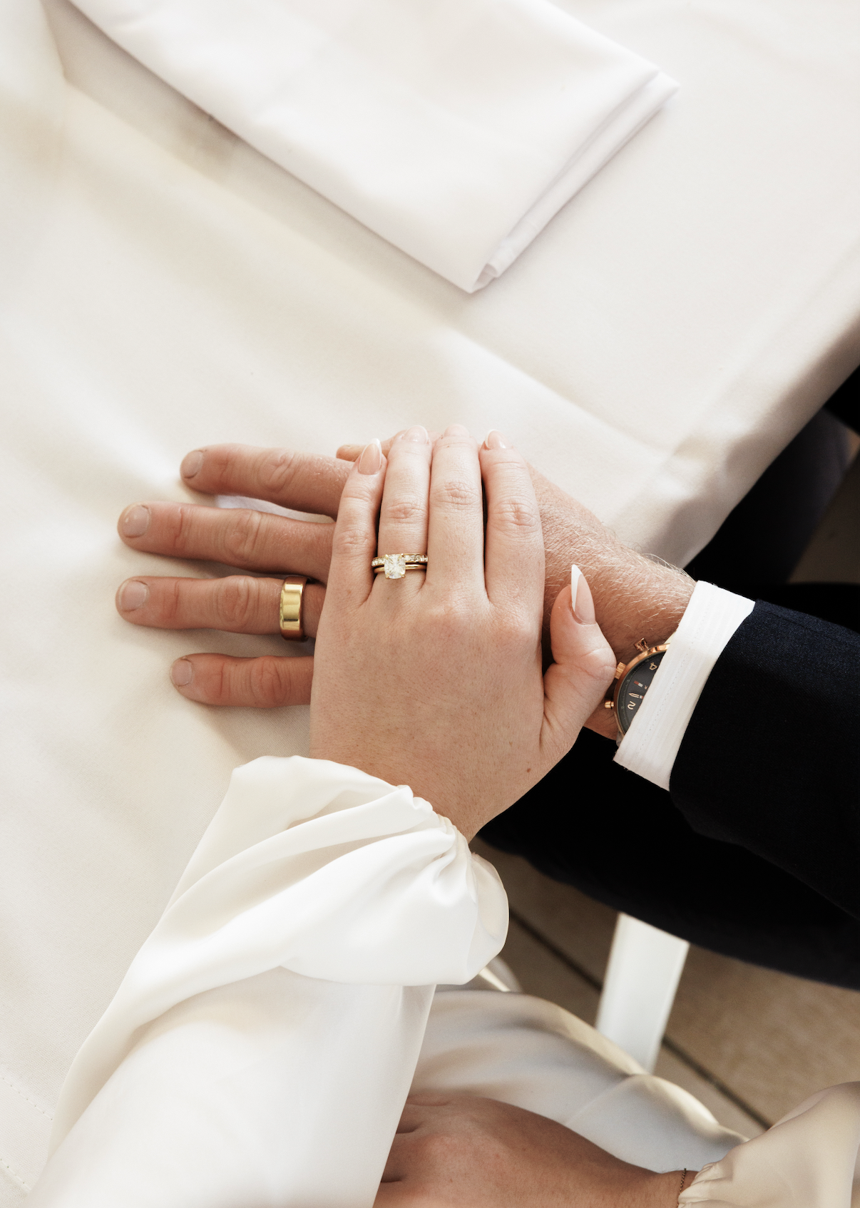 Close-up of a bride and groom's hands resting on a white table. The bride's hand is on top, showing an engagement ring and wedding band. The bridesmaid is wearing a white silk blouse, and the groom is in a dark suit with a watch visible on his wrist.