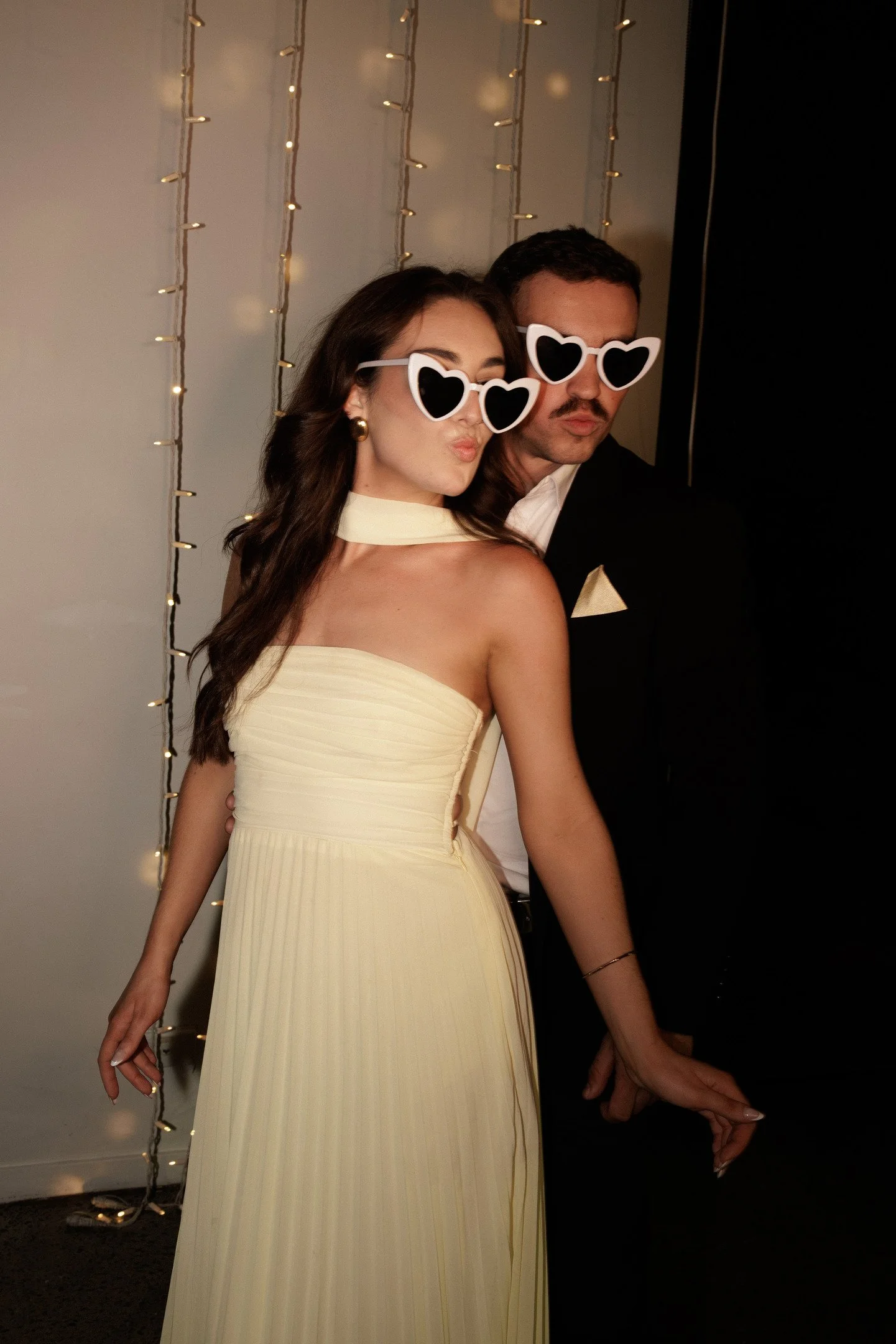 Two people are posing together wearing oversized white sunglasses with heart-shaped black lenses. The woman is dressed in a strapless, light yellow gown with a pleated skirt and is standing close to a man dressed in a black tuxedo with a white shirt 