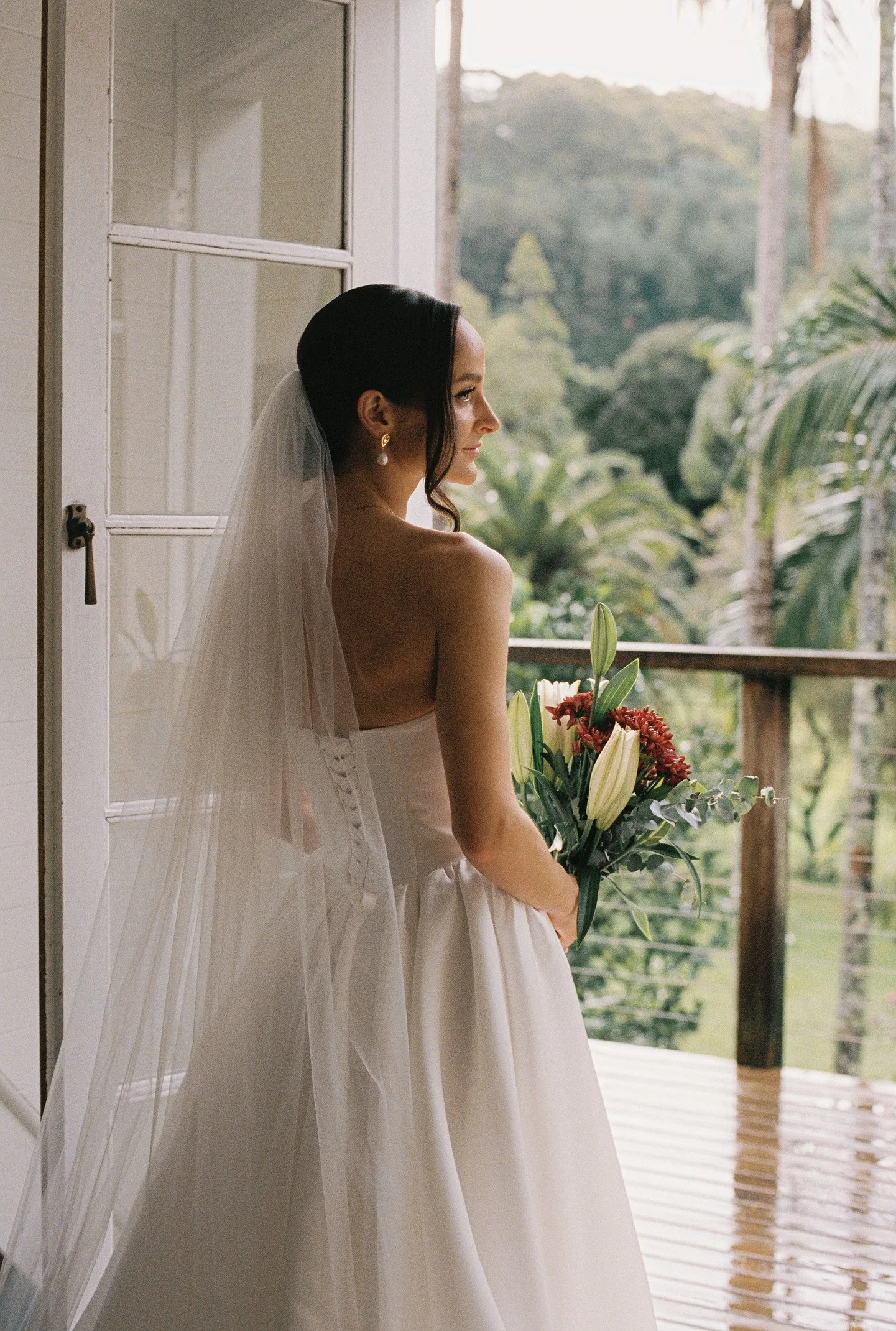 A bride stands on a balcony holding a bouquet of flowers, with her profile visible as she looks outside to a lush, green landscape.