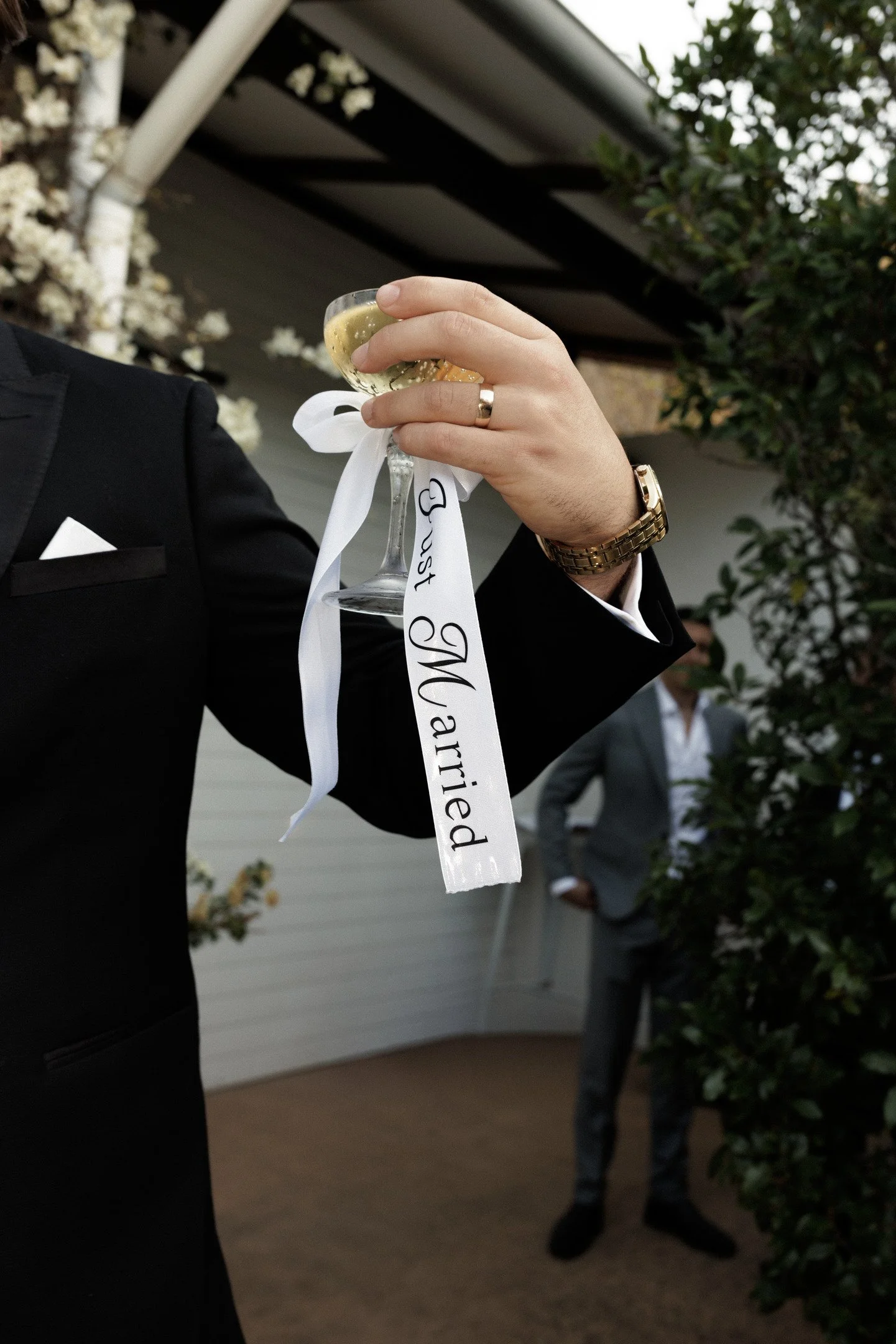 Person in a tuxedo holding a glass of champagne with a white ribbon attached that says 'Just Married', outside near greenery.