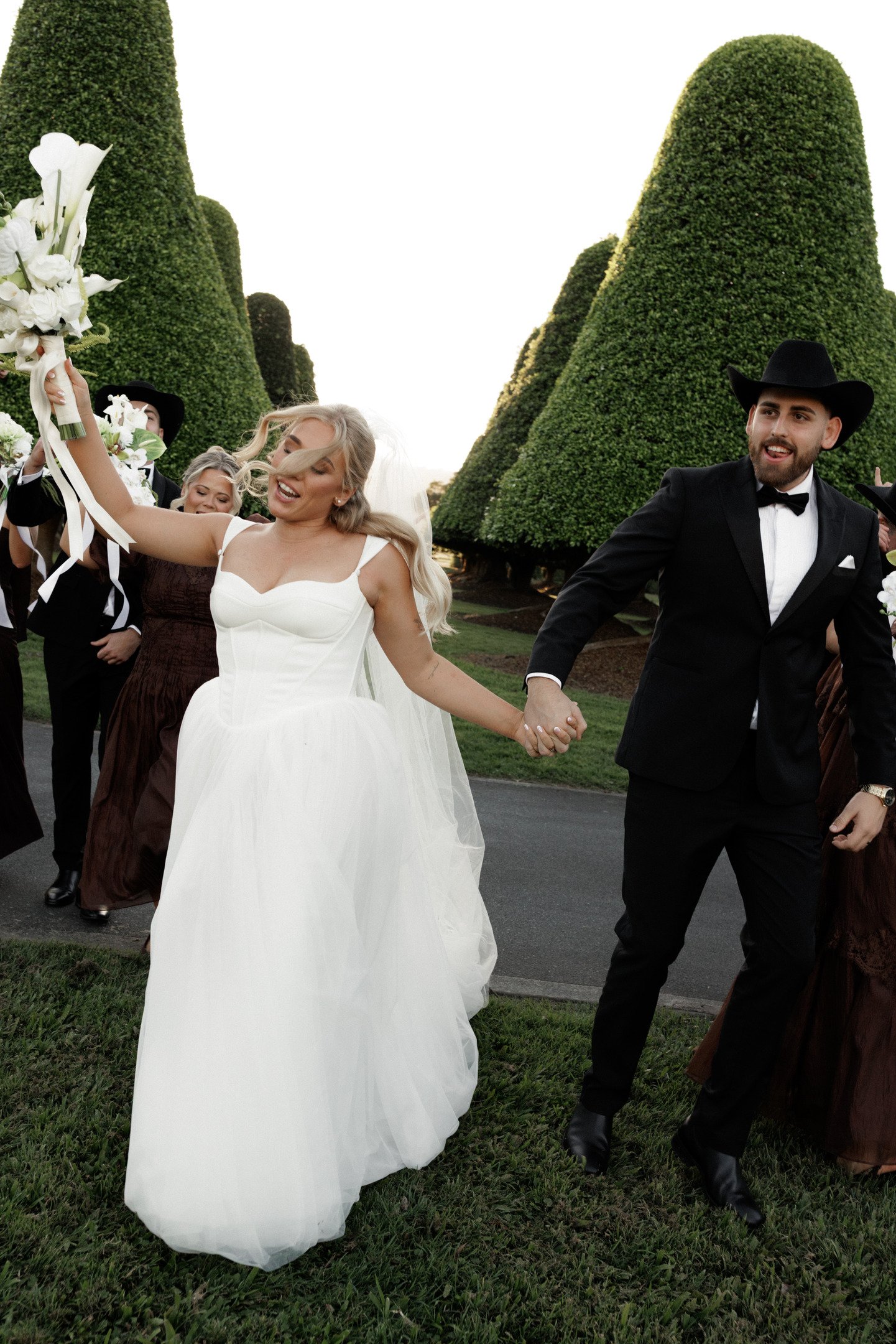 A bride in a white wedding dress holding hands and dancing with a groom in a black tuxedo and black hat outdoors, surrounded by other wedding guests, with tall green trees in the background.