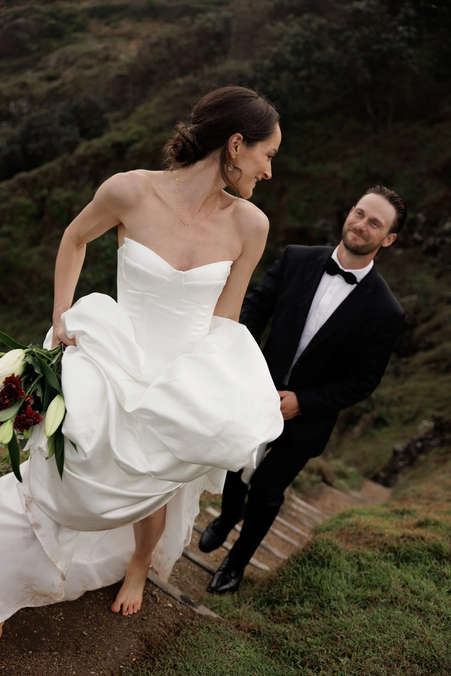 A bride in a strapless white wedding dress and barefoot holding a bouquet of flowers, smiling at a groom in a black tuxedo with a bow tie, outdoors with a grassy hillside background.