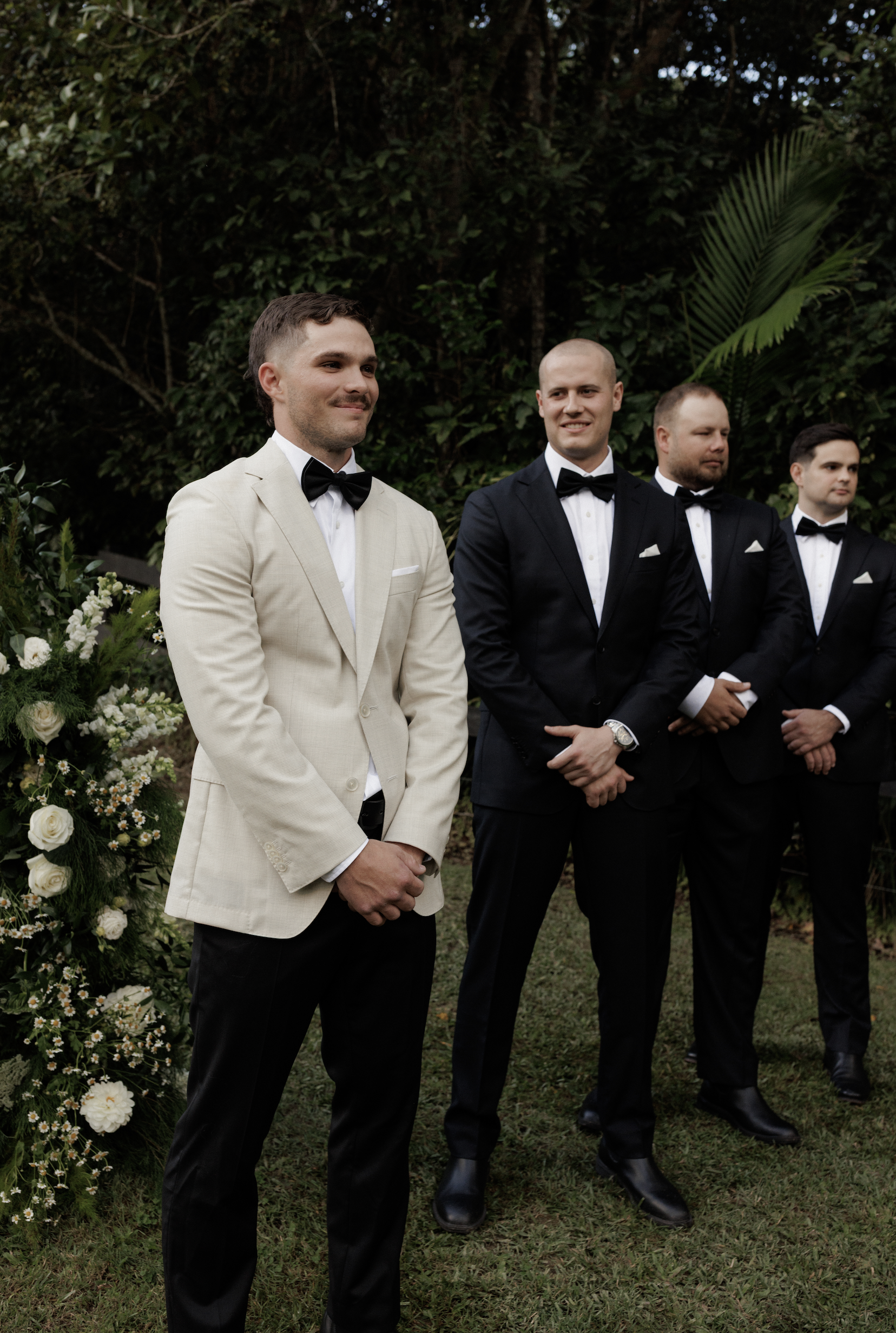 Four men dressed in tuxedos, standing outdoors in front of a greenery backdrop, during a formal event or wedding ceremony.