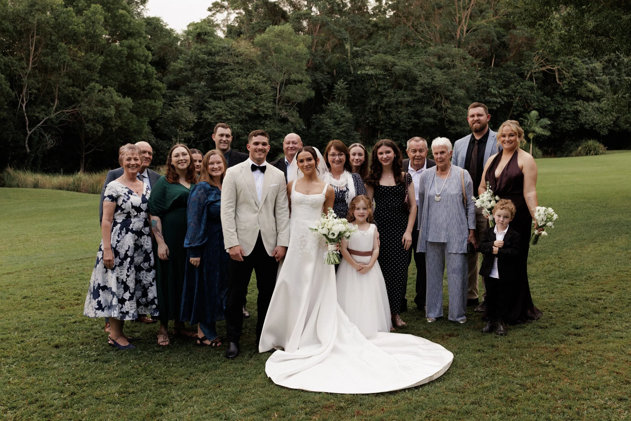 A large group of people, including a bride and groom, posing outdoors on a grassy field with trees in the background. The bride is wearing a white wedding dress and holding a bouquet. The groom is wearing a white tuxedo jacket and black bow tie. The 