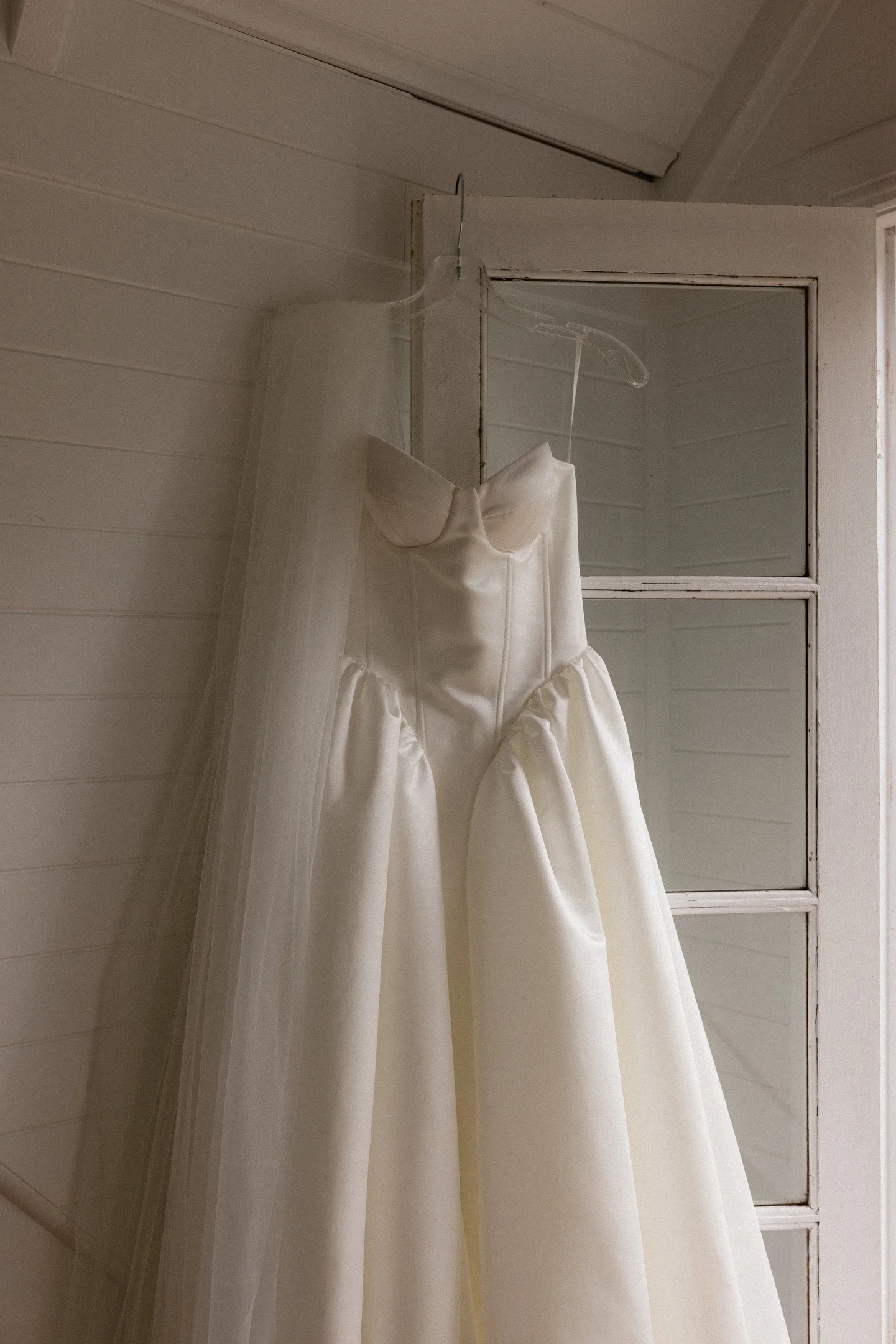 A white wedding dress hanging on a clear plastic hanger attached to a window latch, with a sheer white veil draped over the dress.