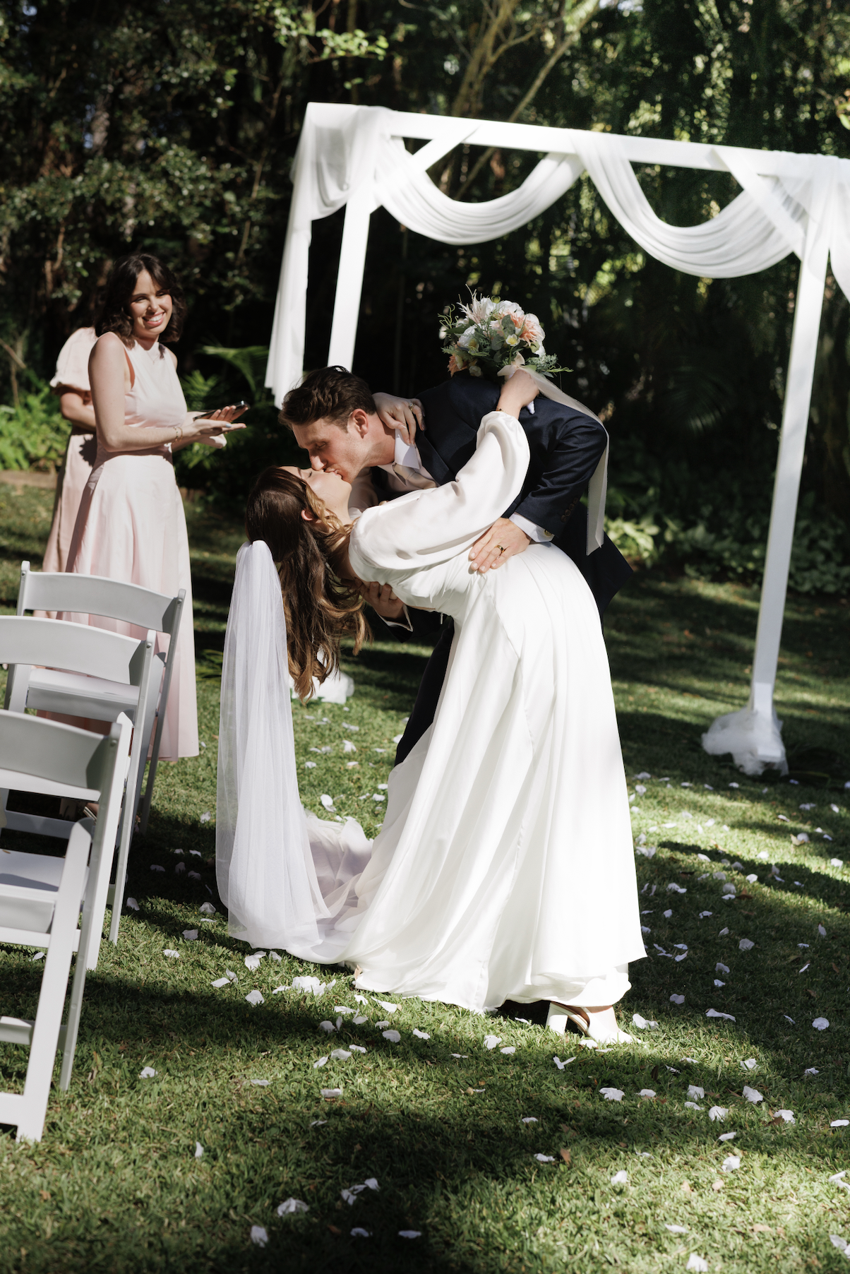 A bride and groom share a kiss at an outdoor wedding ceremony, with a woman in a pink dress smiling nearby and a decorative white arch with draped fabric behind them.