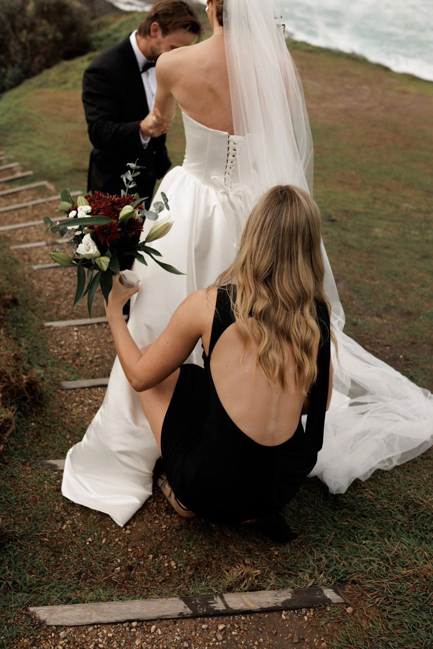 A wedding ceremony on the beach with a bride, groom, and a woman in black dress holding a bouquet of flowers.