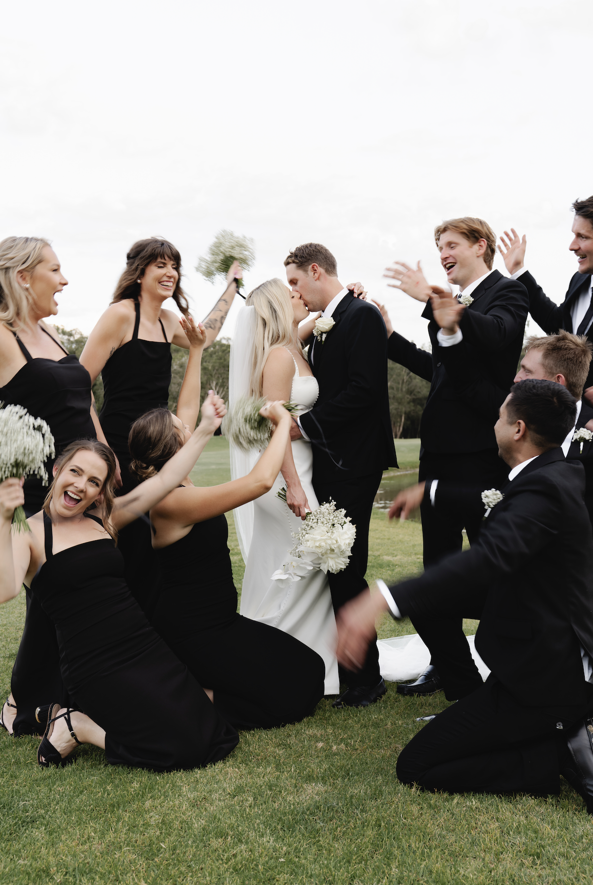 Happy wedding party celebrating outdoors, with the couple kissing in the center, surrounded by bridesmaids and groomsmen cheering and holding bouquets on a grassy field under an overcast sky.