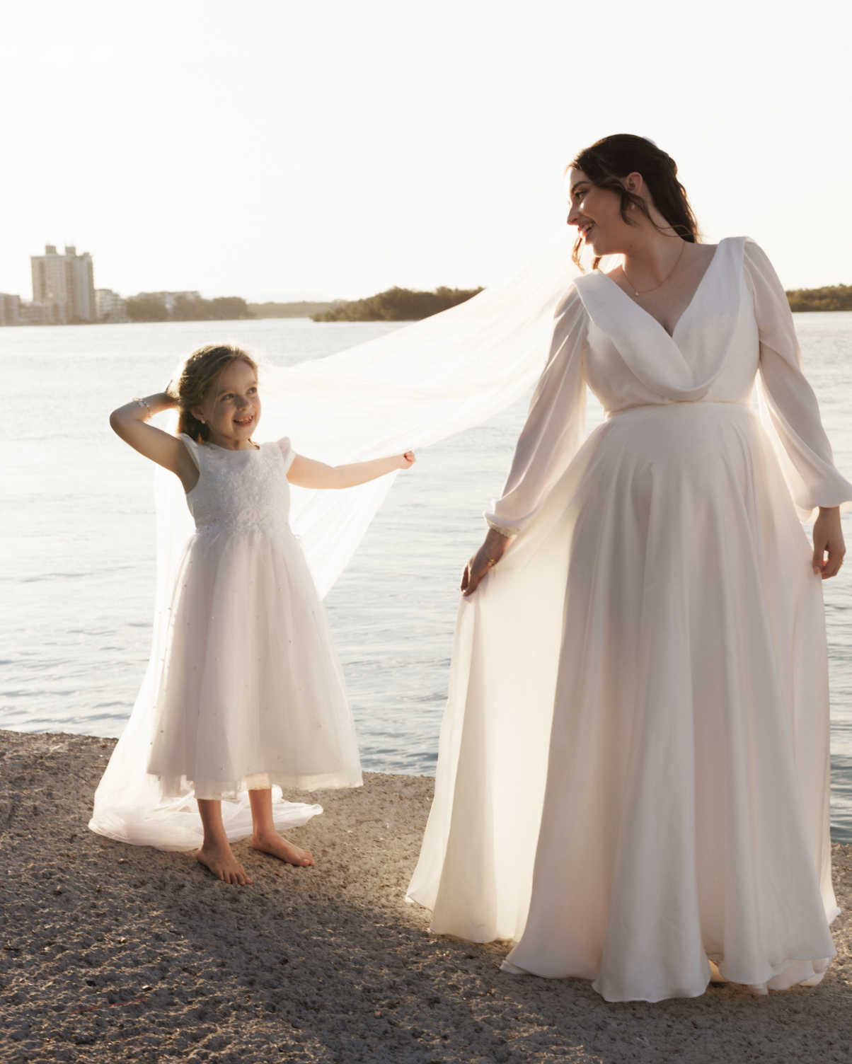 A woman and a young girl, both dressed in white dresses, standing on a rocky shoreline near a body of water during sunset, smiling at each other.