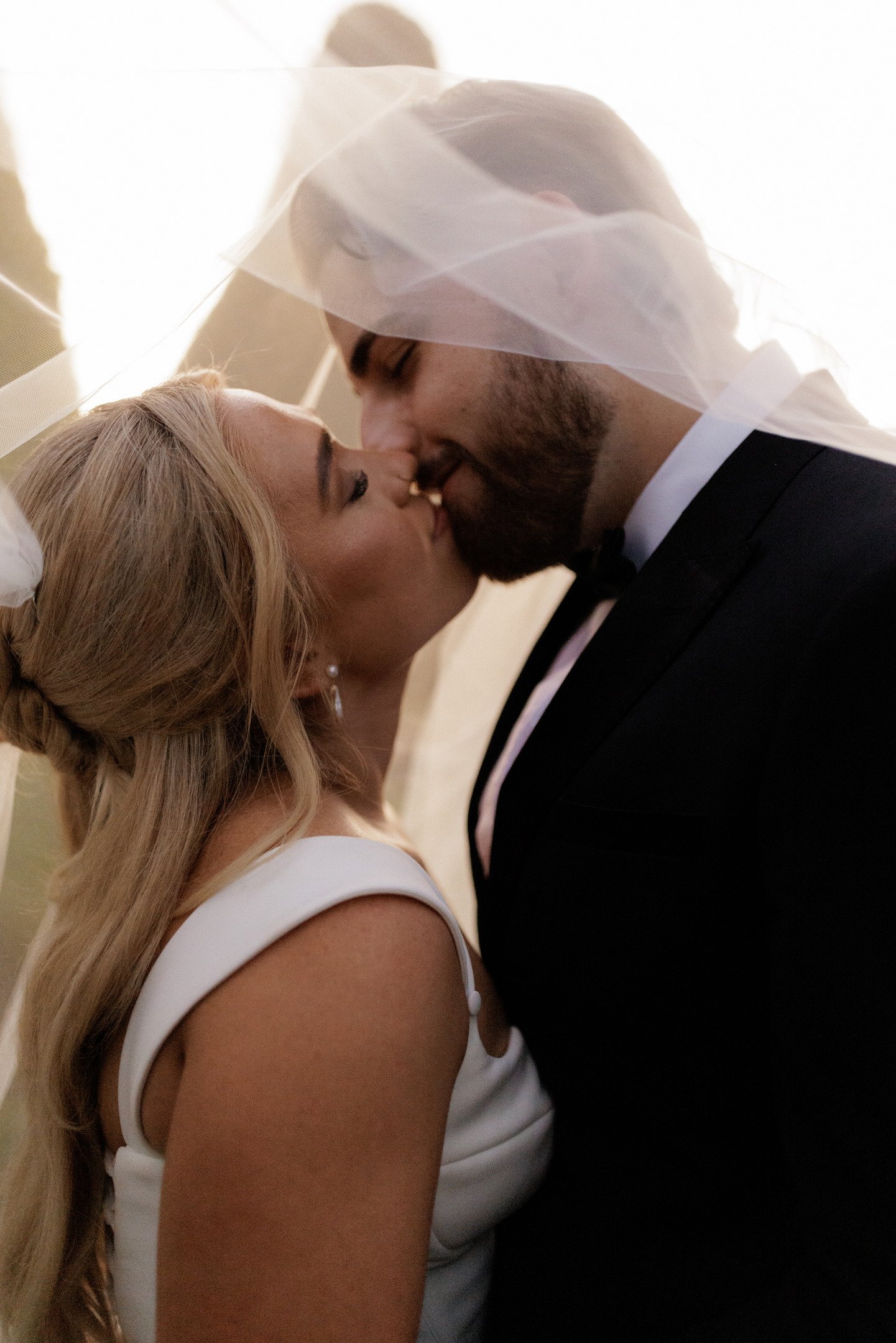 A couple dressed in formal wedding attire, about to kiss, with the woman wearing a white dress and veil, and the man wearing a black tuxedo with a bow tie.