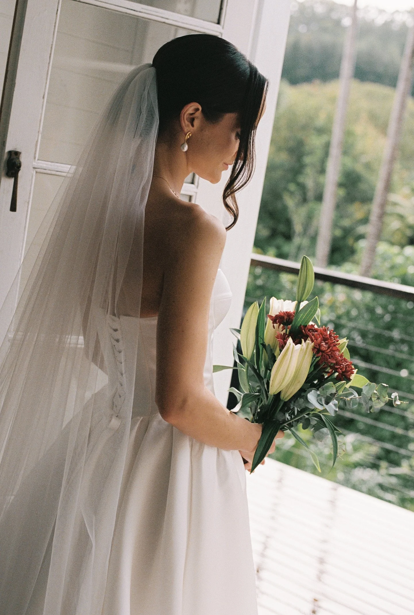 A bride in a white wedding gown holding a bouquet of flowers, standing near a porch with a view of greenery outside.