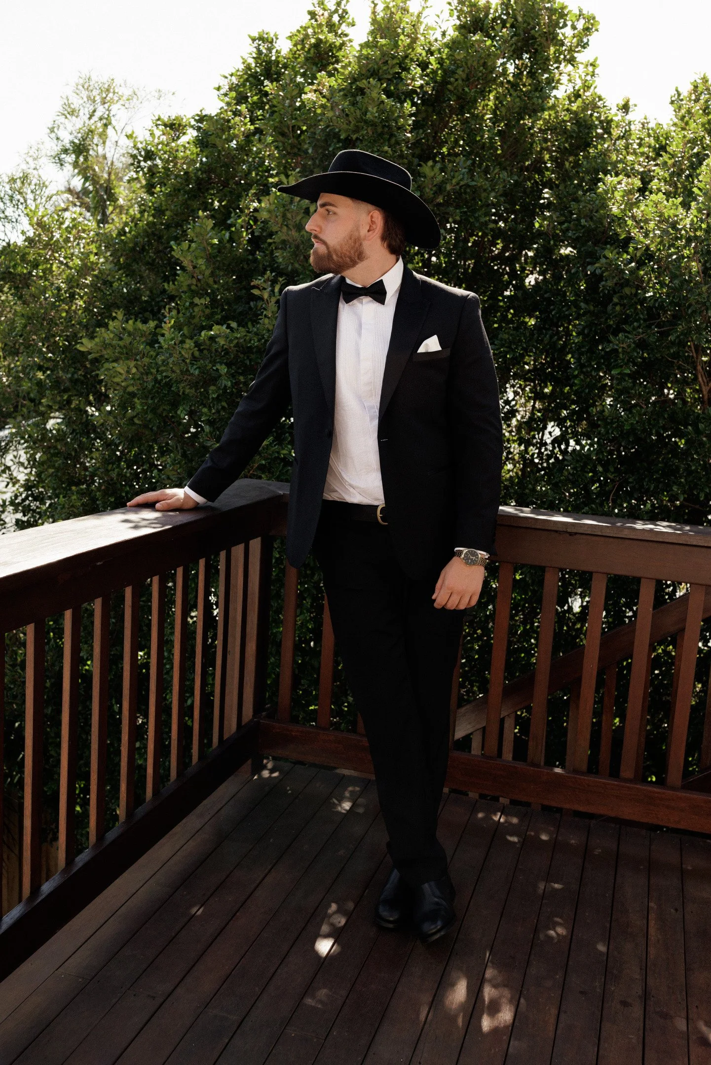 A man dressed in a black tuxedo, white shirt, and black bow tie, wearing a wide-brimmed black hat, standing on a wooden balcony with greenery in the background.