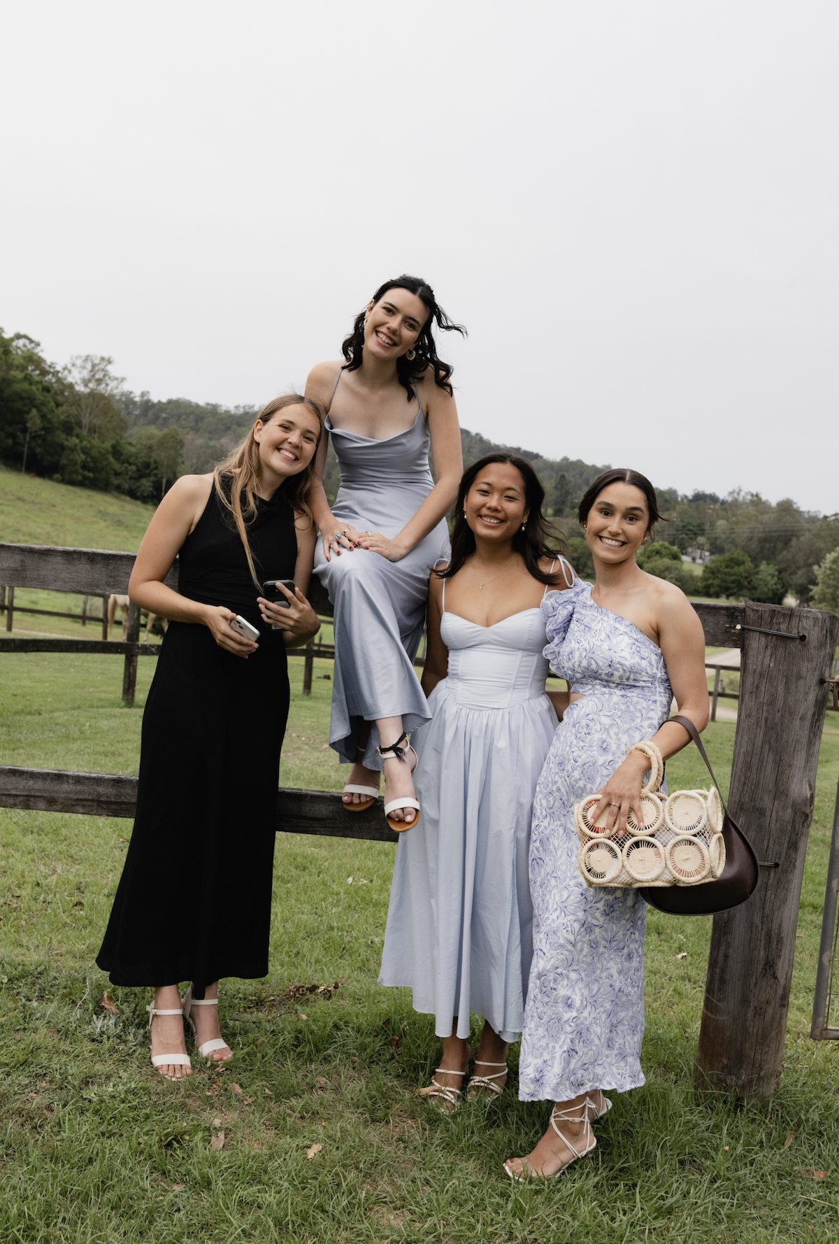 Five women smiling outdoors in a grassy field with a wooden fence and hills in the background. Four are standing, one is sitting on the fence.