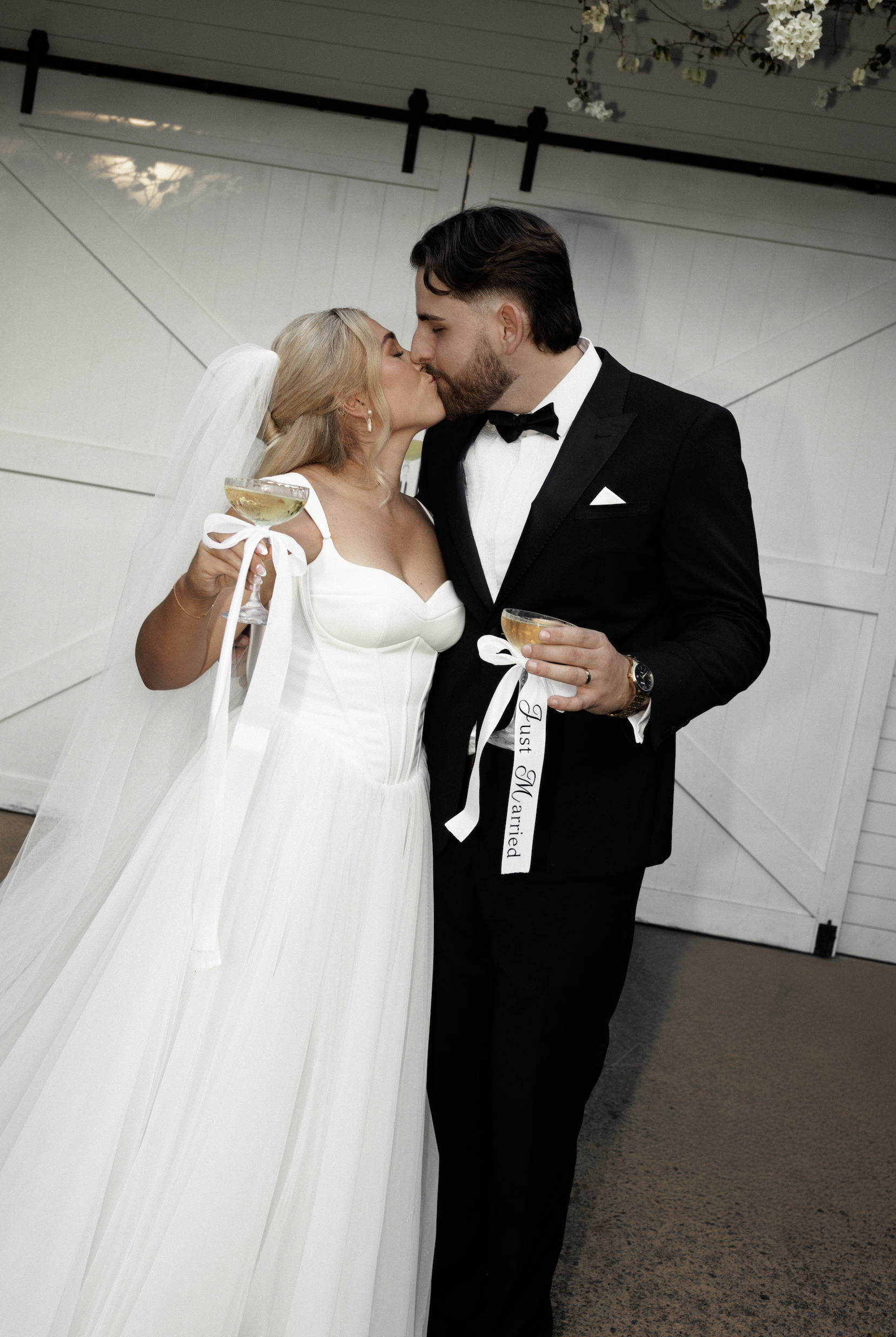 A bride and groom sharing a kiss at their wedding, holding champagne glasses with ribbons, in front of a white barn-style background.