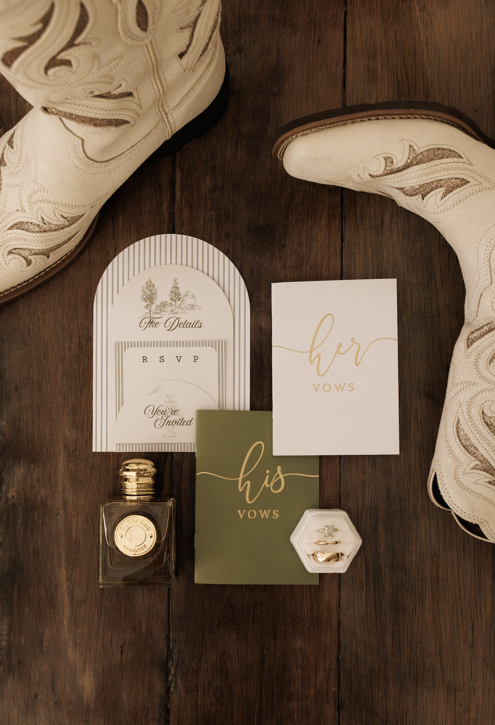 Wedding invitation cards, a small perfume bottle, and two rings in a ring box arranged on a wooden surface, along with a pair of white boots with gold embellishments.