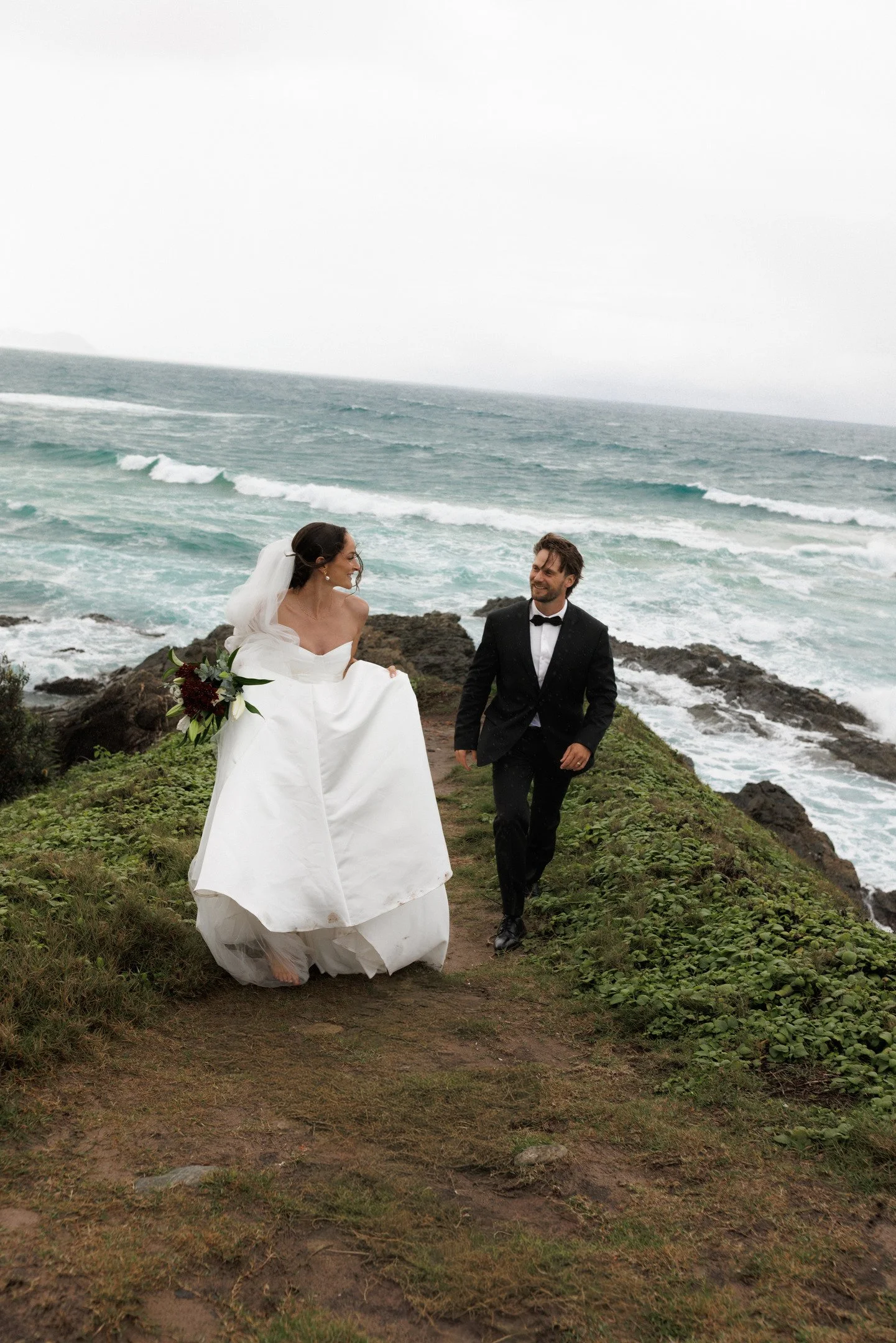 A bride and groom walking along a rocky path by the ocean, with waves and a cloudy sky in the background. The bride is wearing a white wedding dress and veil, holding a bouquet, and the groom is dressed in a black tuxedo with a bow tie.
