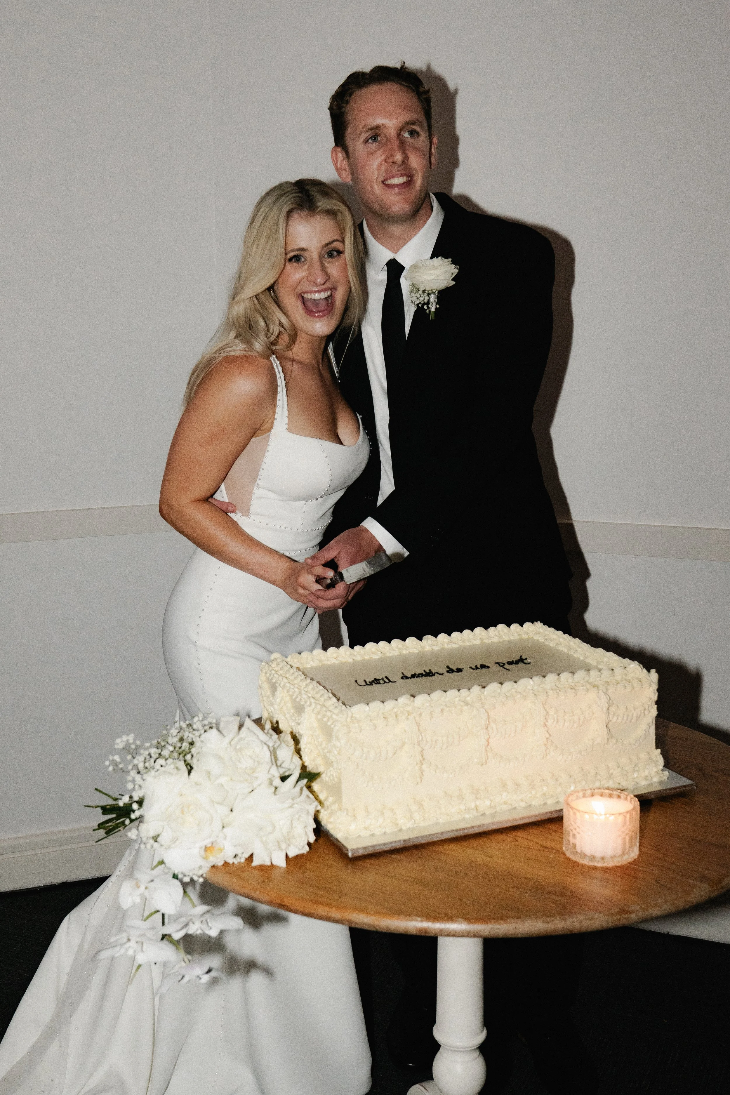 A bride and groom celebrating their wedding, cutting a cake together, with a bouquet of white flowers and a lit candle on the table.