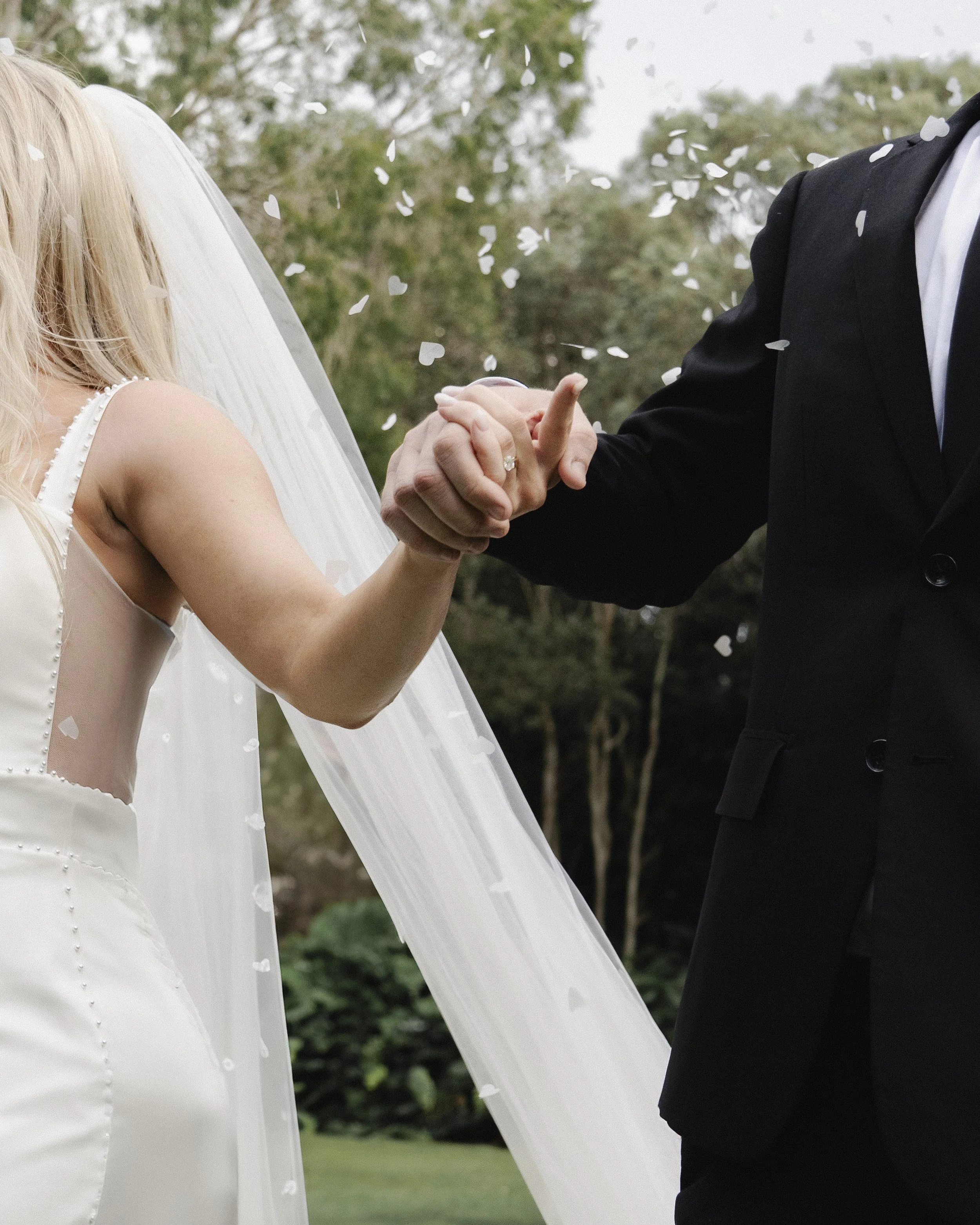 A bride and groom holding hands during their wedding ceremony outside, surrounded by greenery, with white confetti falling around them.