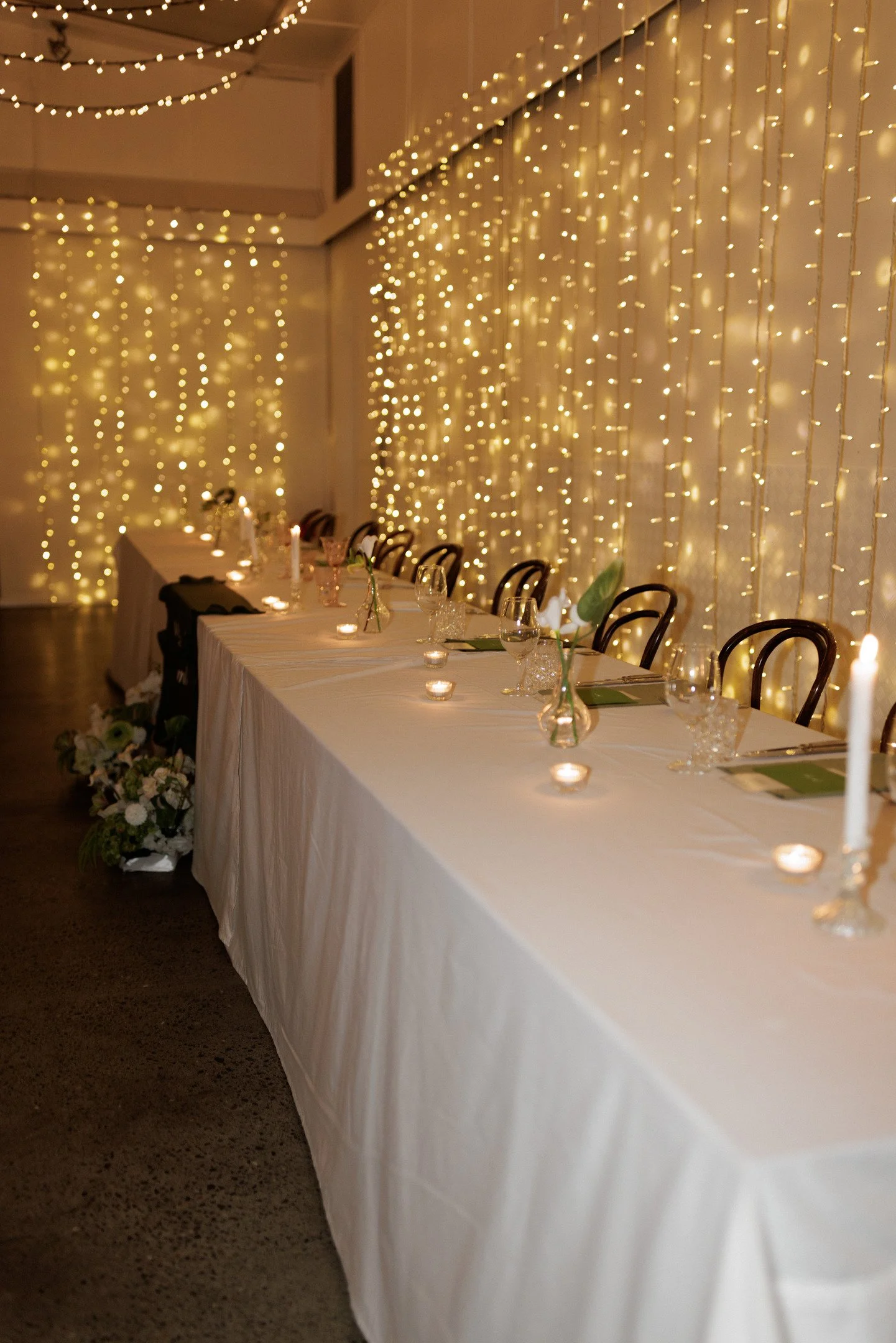 A long banquet table decorated with candles and small flowers, set for an event, against a wall with hanging string lights creating a warm ambiance.
