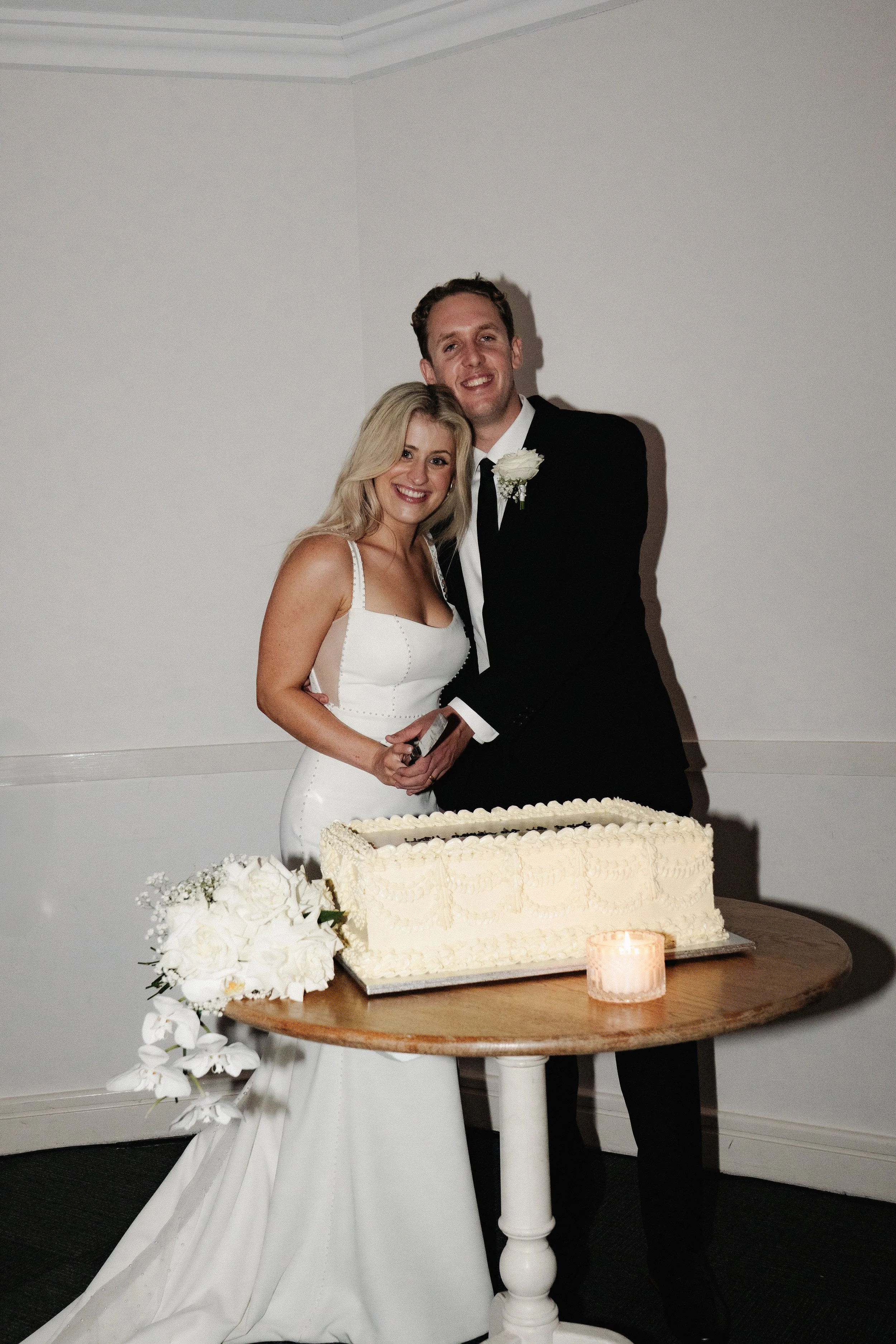 A bride and groom standing together behind a wedding cake on a round wooden table, with a bouquet of white flowers and a lit candle on the table, during their wedding celebration.