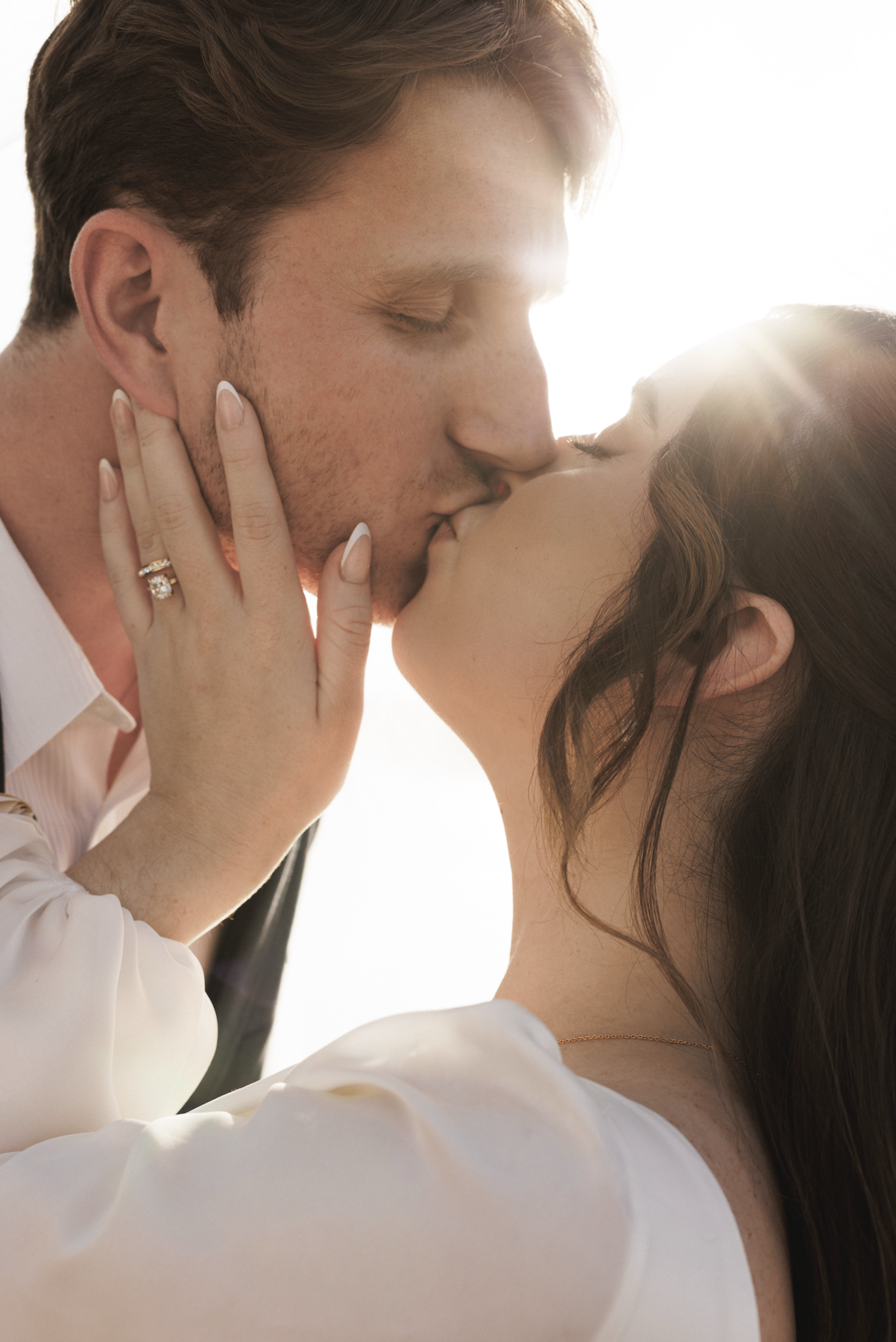 A couple sharing a kiss with sunlight in the background, highlighting their facial features and intimate moment.