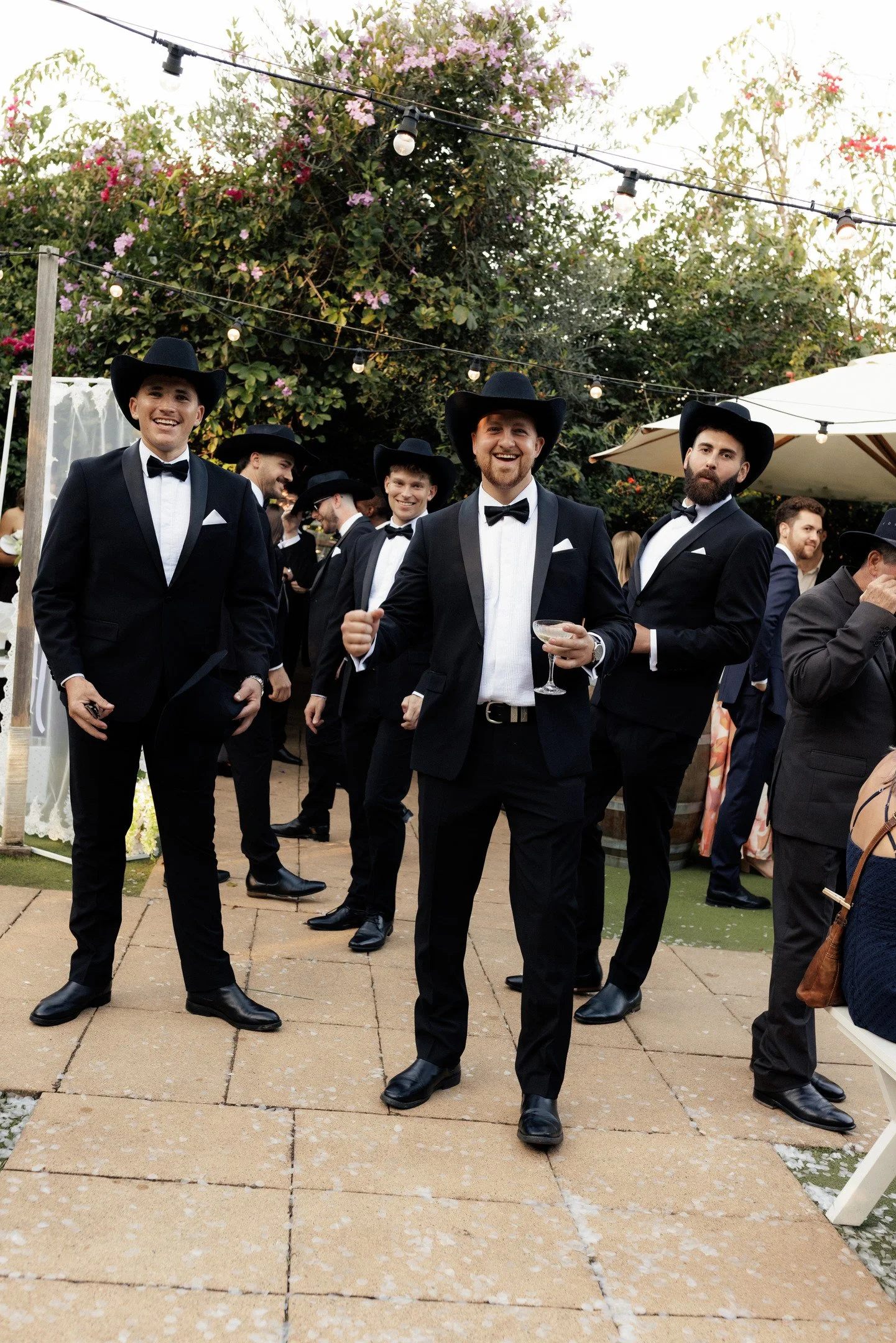 Group of men in tuxedos and cowboy hats at an outdoor event with string lights and floral decorations.
