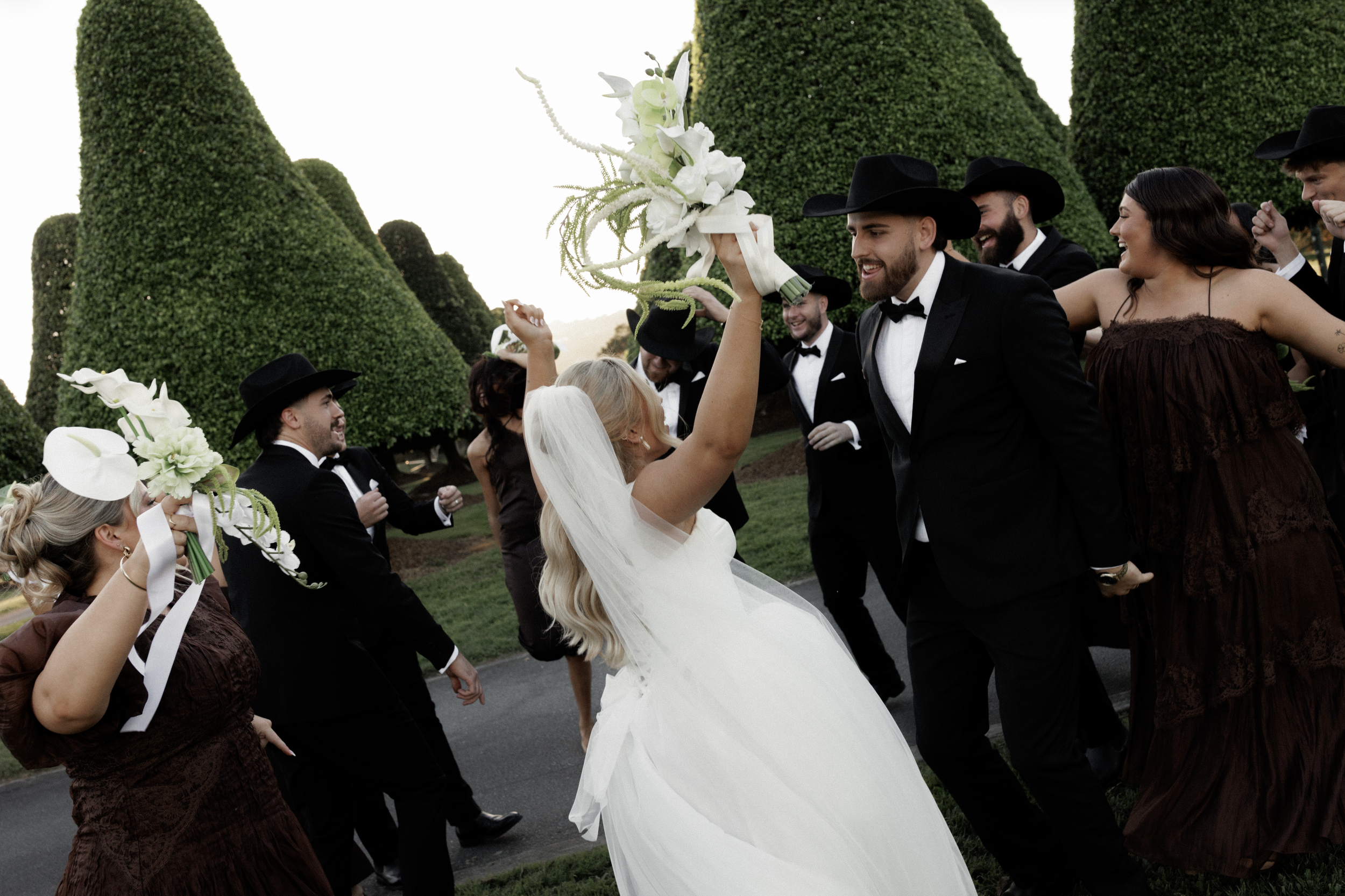 A bride in a white wedding dress and veil is holding a bouquet of white flowers, surrounded by guests in western attire and hats, outdoors with large green bushes in the background.