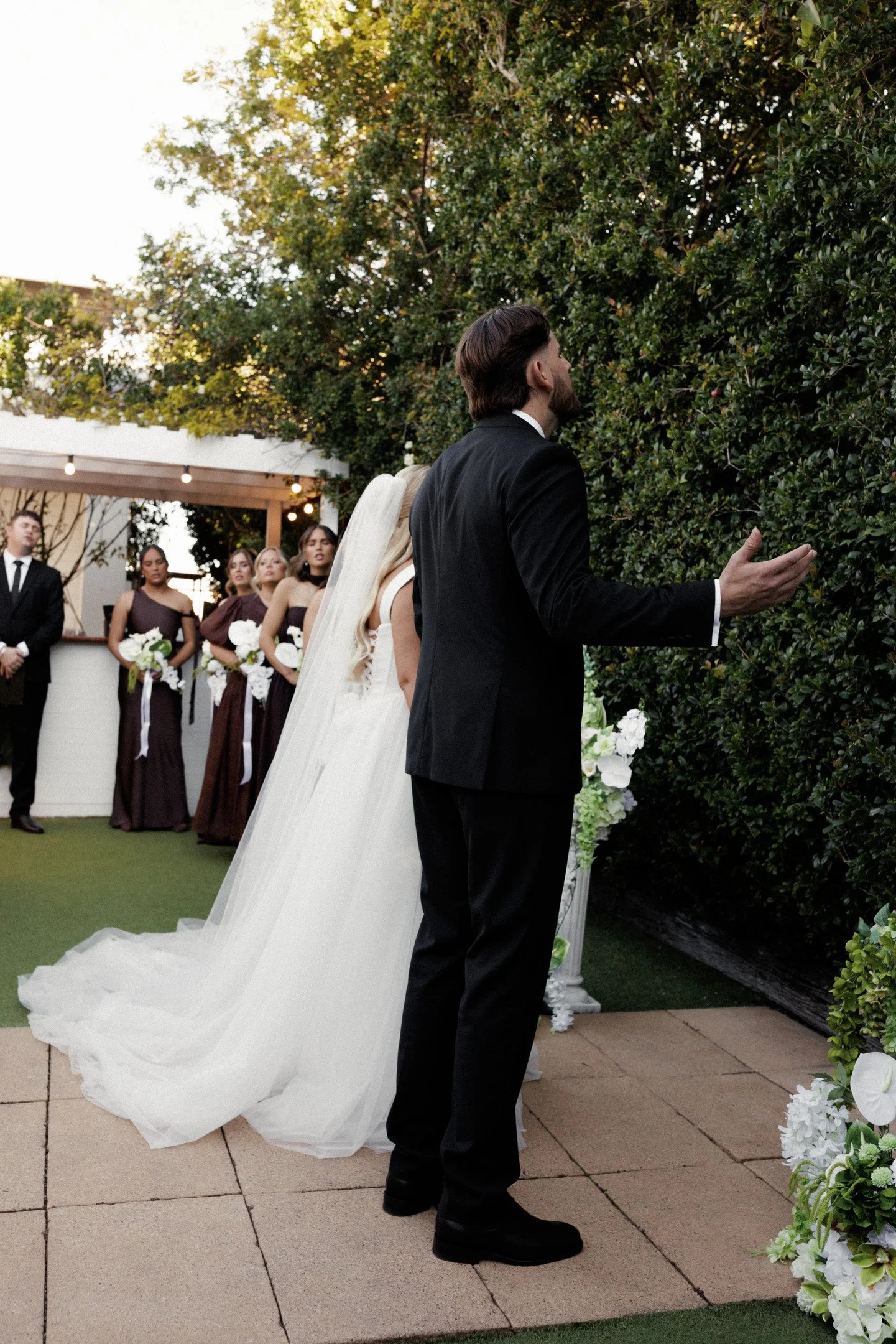 A groom in a black tuxedo stands facing a bride in a white wedding dress with a long train, during an outdoor wedding ceremony. The groom is reaching out towards a green hedge, which is decorated with white flowers. Bridesmaids in purple dresses and 